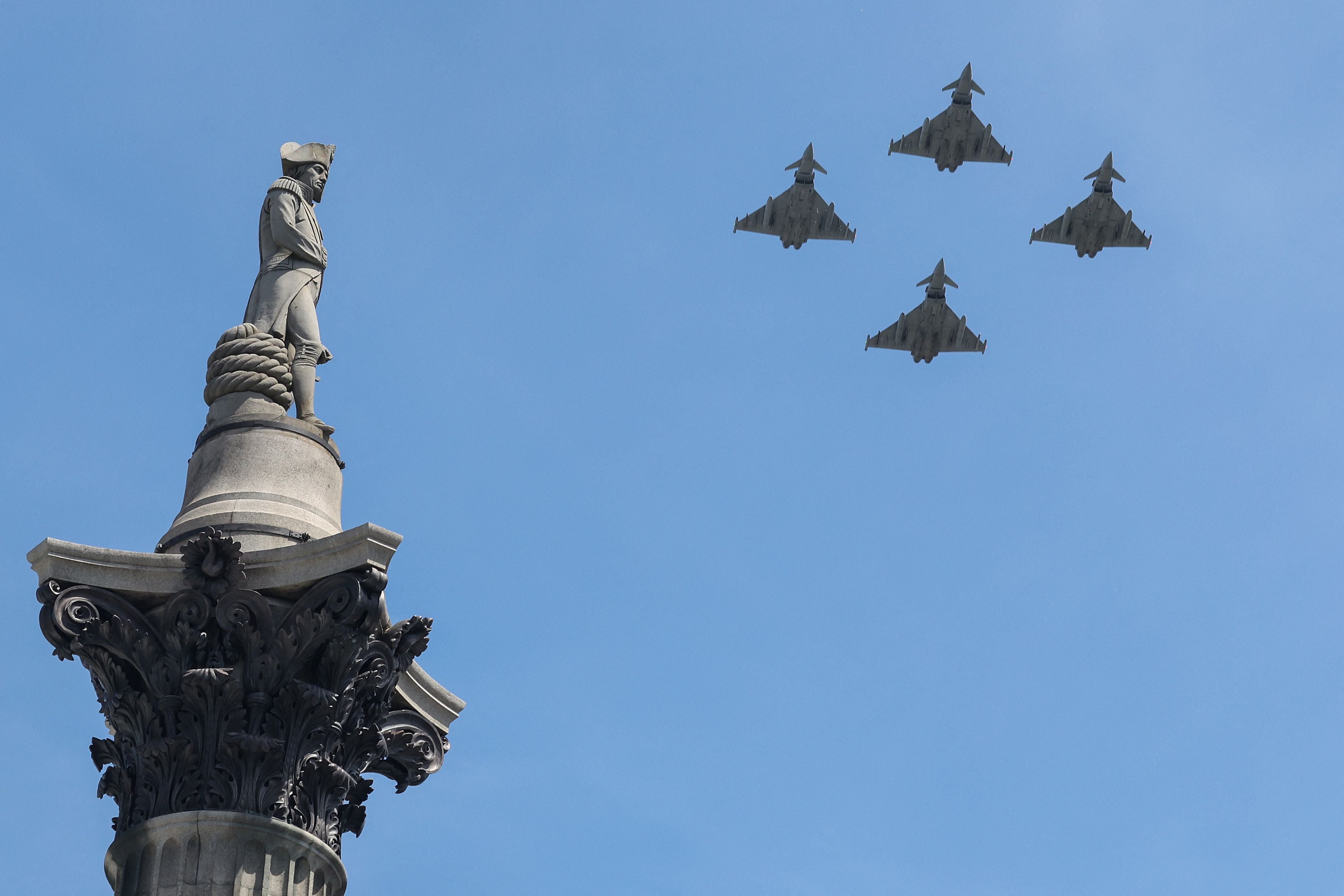 Vier Kampfjets fliegen in Formation neben einer Statue auf einer Säule vor blauem Himmel.