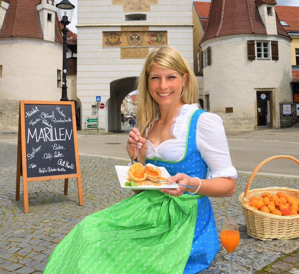 Frau in Dirndl isst Marillenknödel, Korb mit Marillen und Glas Saft daneben, Altstadt im Hintergrund.