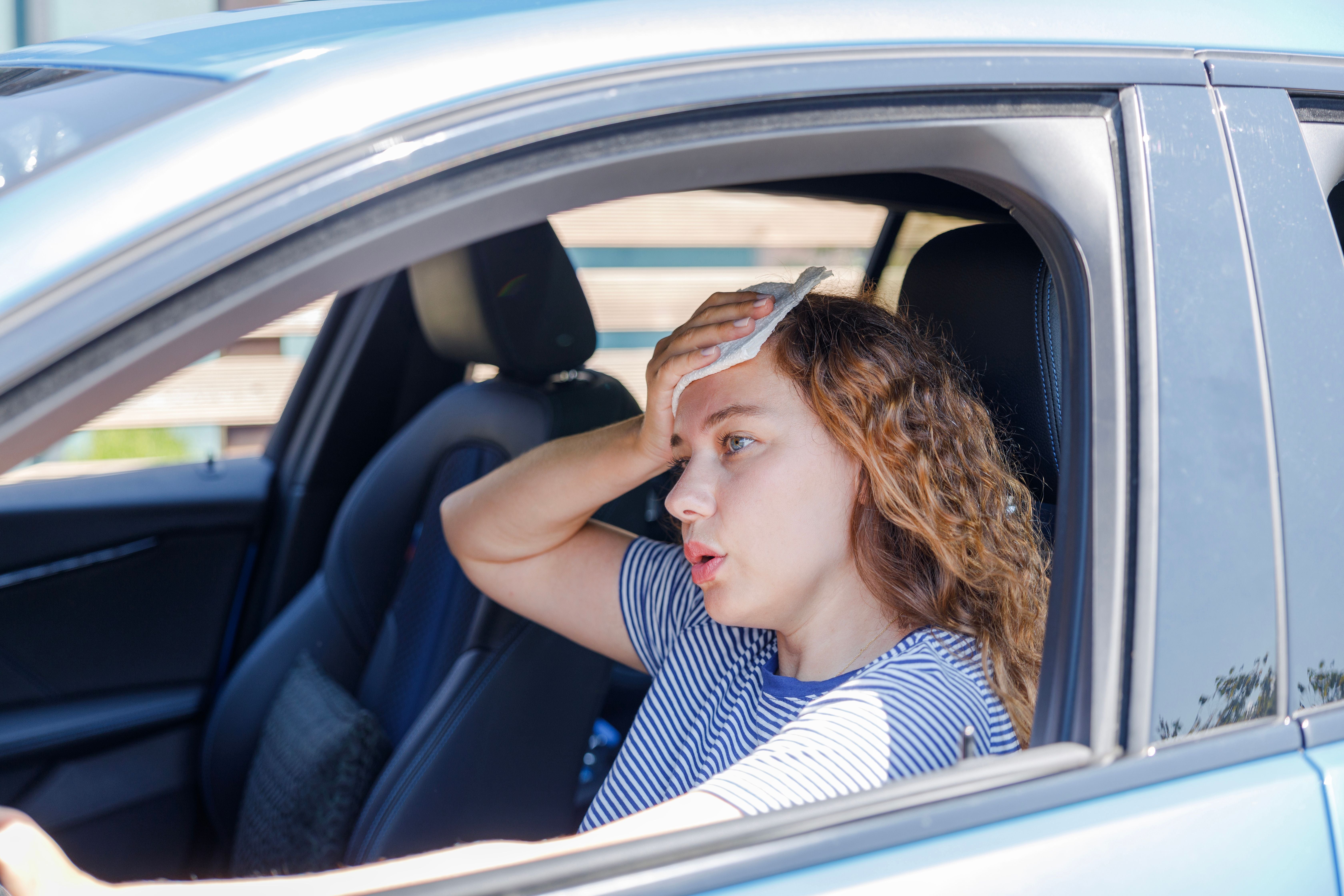 Junge Frau sitzt im Auto und wischt sich wegen extremer Hitze den Schweiß von der Stirn.
