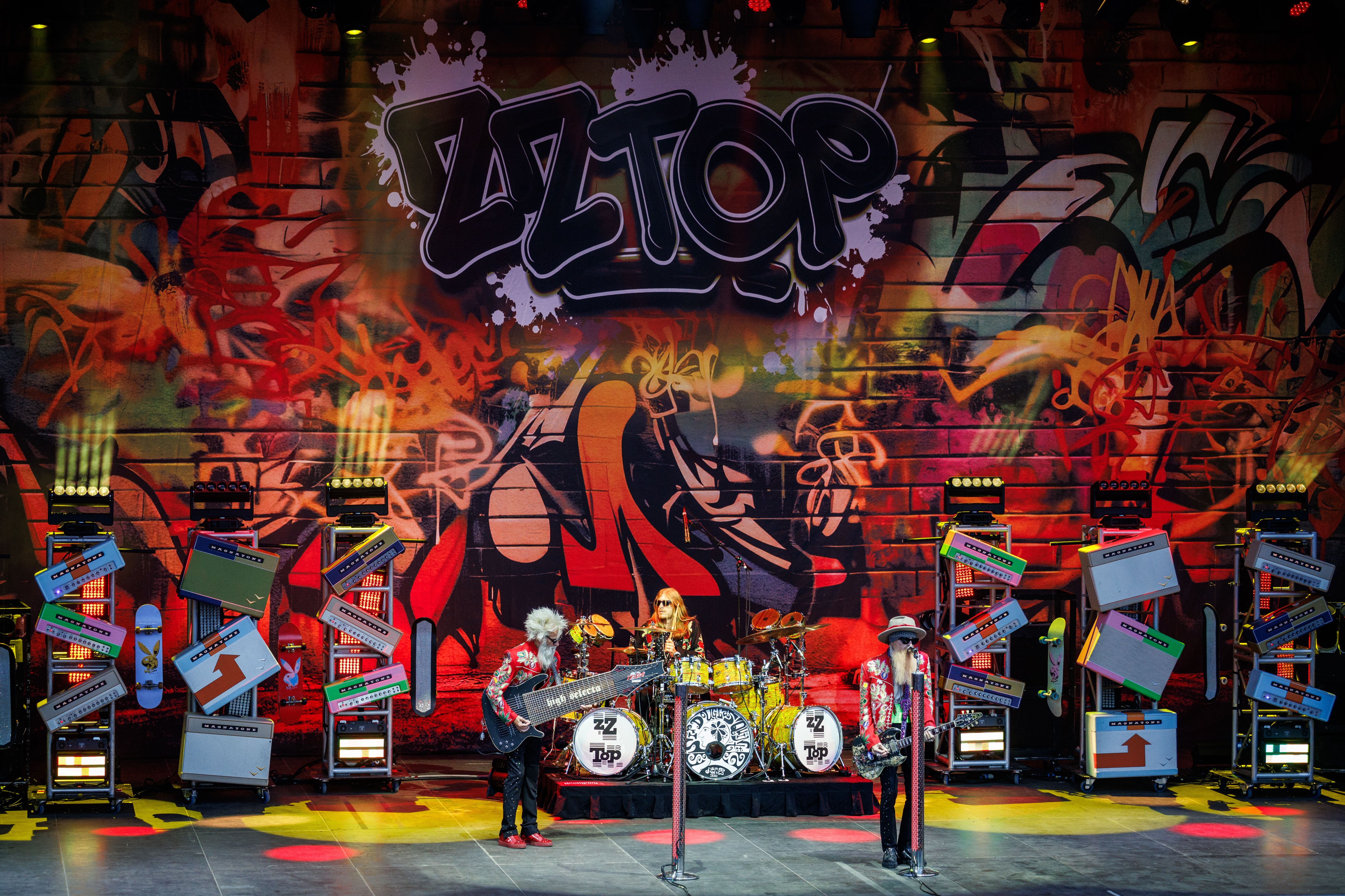 Die Band ZZ Top spielt auf einer bunt beleuchteten Bühne mit Graffiti-Hintergrund.