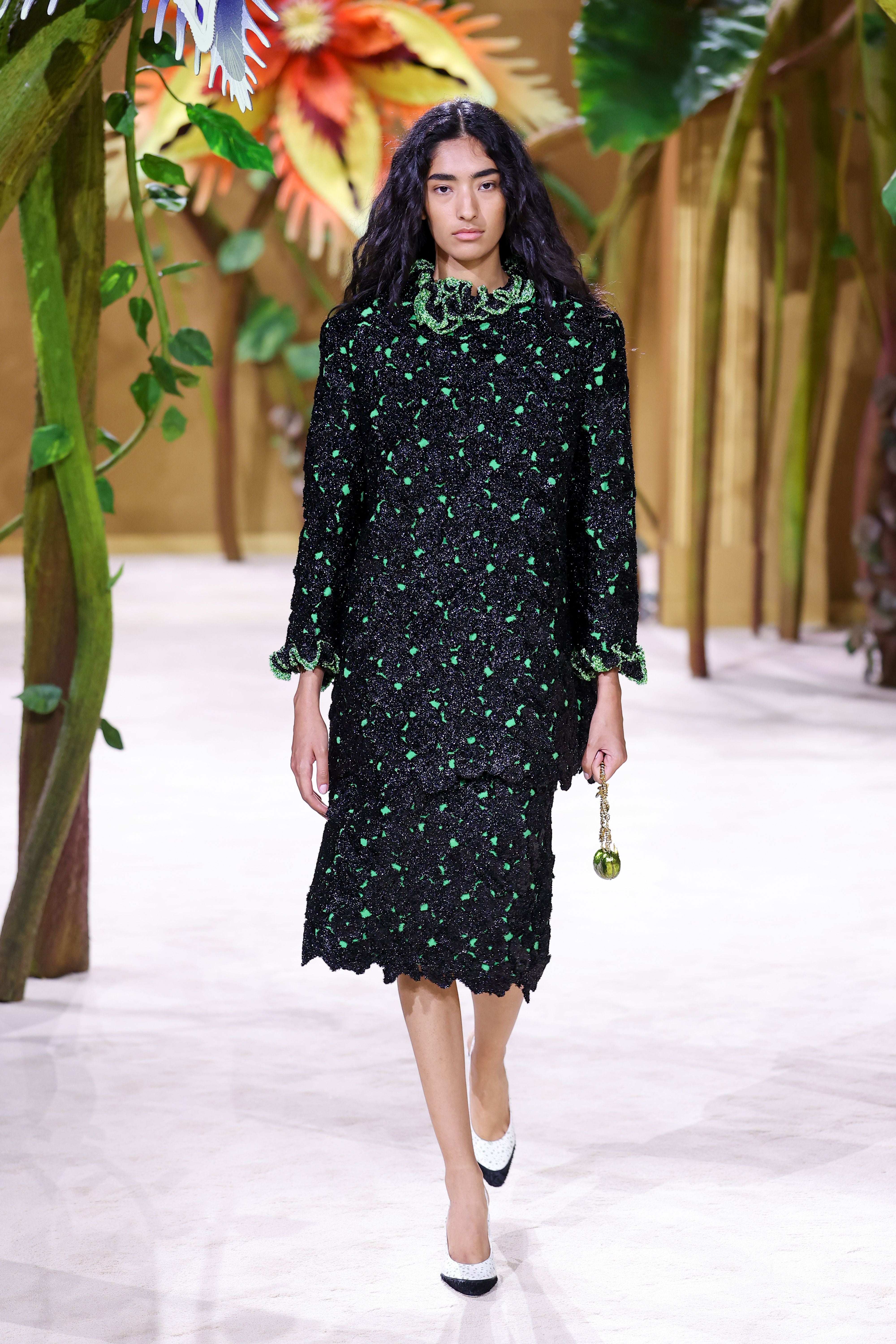 Model in schwarzem Kleid mit grünen Details auf dem Laufsteg, umgeben von großen Pflanzen.