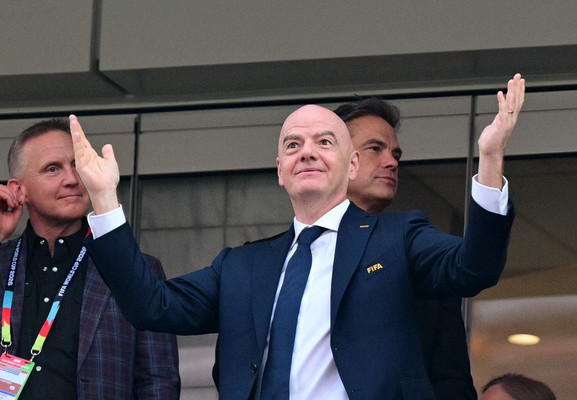 Ein Mann im Anzug hebt beide Arme, neben ihm stehen zwei weitere Männer im Stadion.