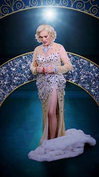 Eine Frau in funkelndem Abendkleid steht auf einer Bühne mit glamourösem Hintergrund.
