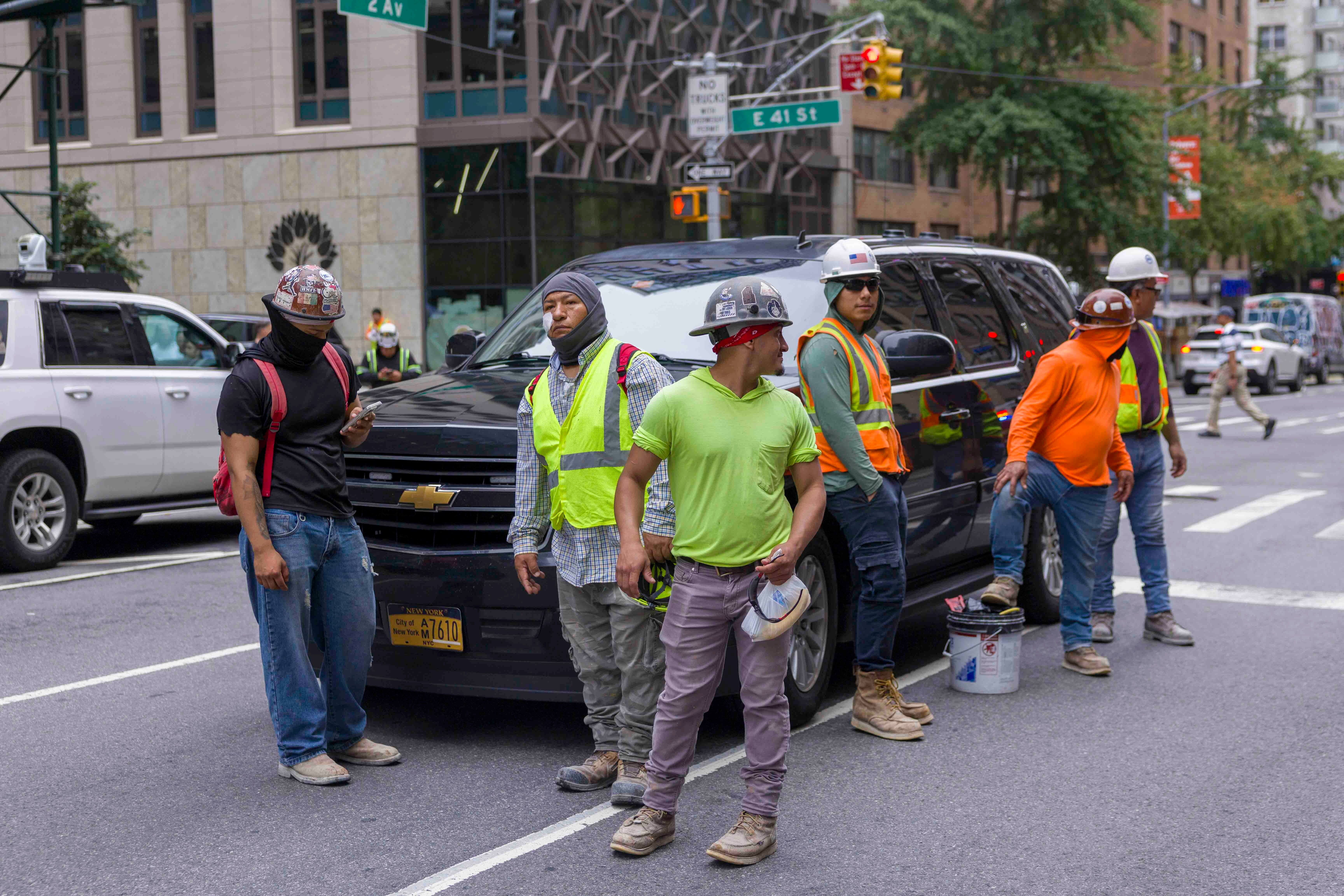Sechs Bauarbeiter mit Helmen stehen vor einem schwarzen SUV in einer Straßenszene in Manhattan.