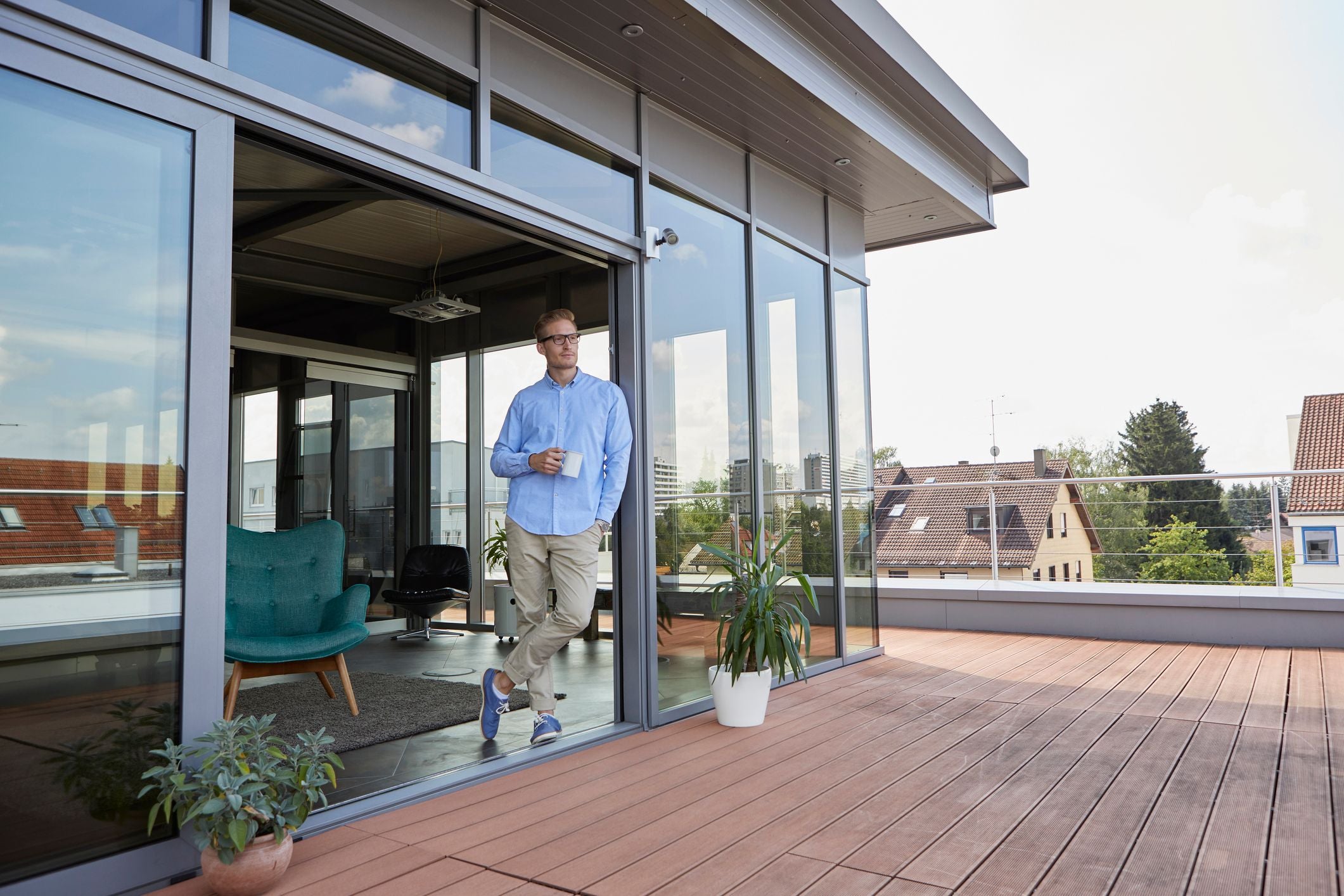 Mann steht mit Kaffeetasse auf moderner Terrasse vor verglastem Wohnzimmer, Pflanzen im Vordergrund.
