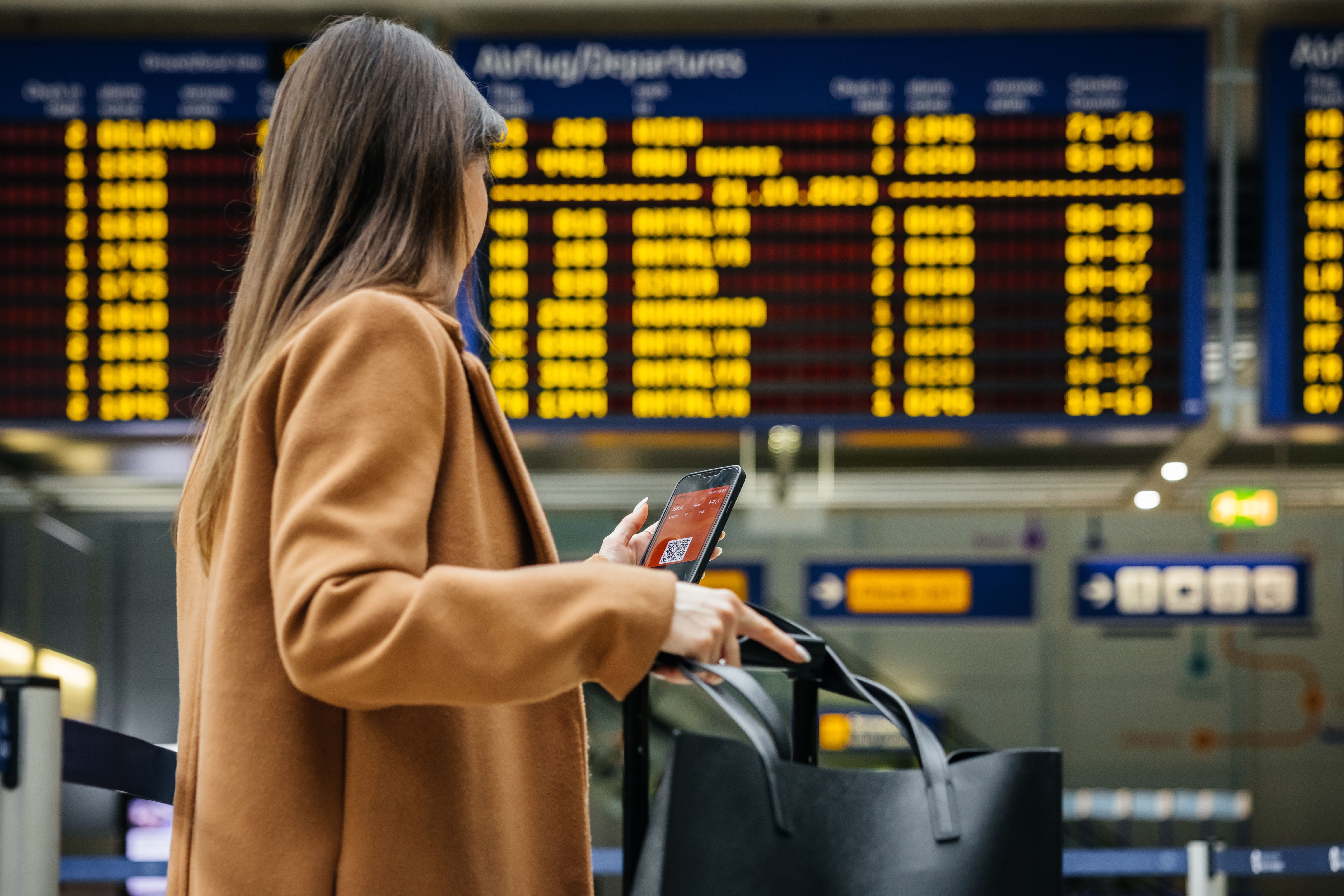 Frau mit Handy steht vor Abflugtafel am Flughafen und hält Gepäck.