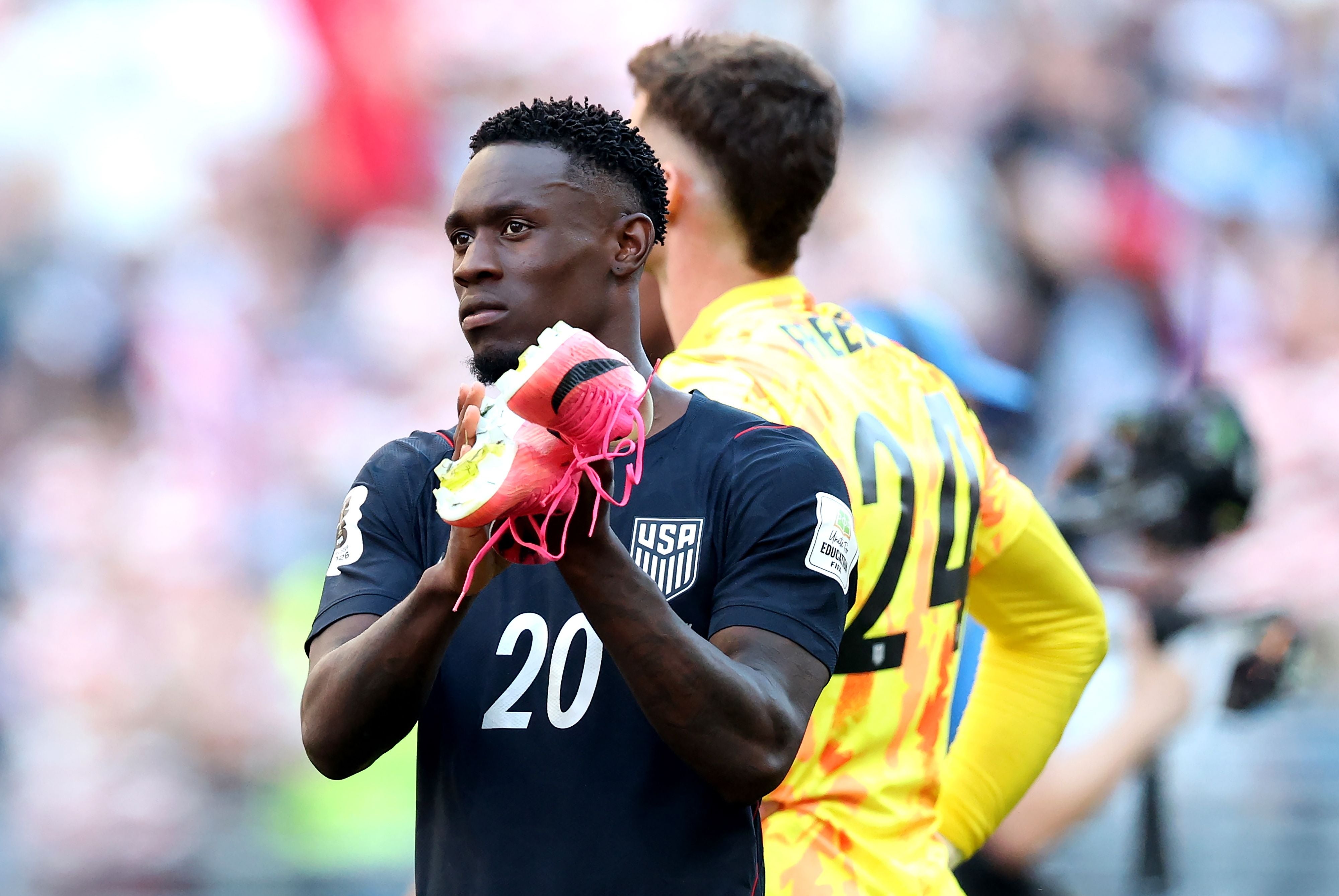 US-Fußballspieler im dunklen Trikot klatscht Beifall und hält Schuhe in der Hand nach einem Spiel.