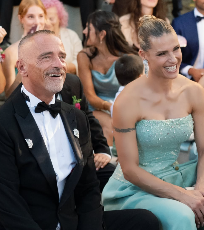 Ein Mann im Smoking und eine Frau im türkisfarbenen Kleid sitzen lachend nebeneinander.