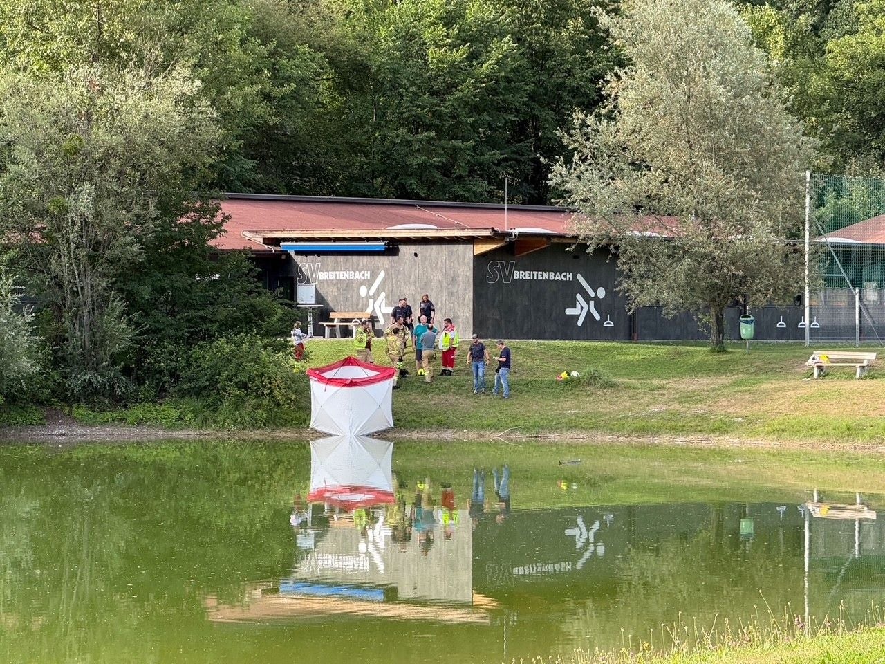Rettungskräfte und Personen stehen am Ufer eines Teichs vor einem Gebäude mit der Aufschrift SV Breitenbach.