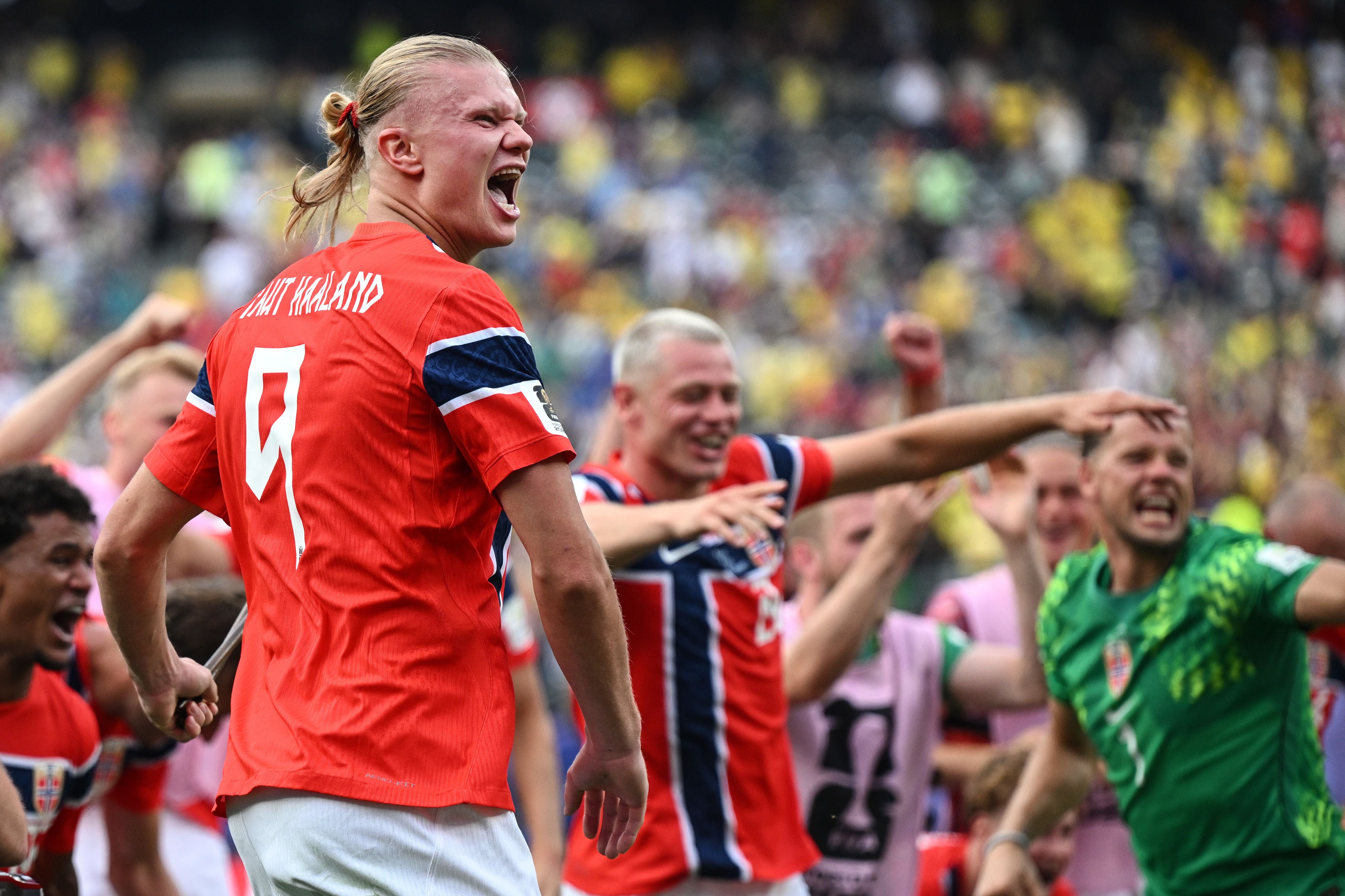 Ein Fußballer mit blondem, zum Zopf gebundenem Haar jubelt lautstark im roten Trikot mit der Rückennummer 9. Um ihn herum feiern weitere Mitspieler in roten und grünen Trikots vor einer vollen Fantribüne.