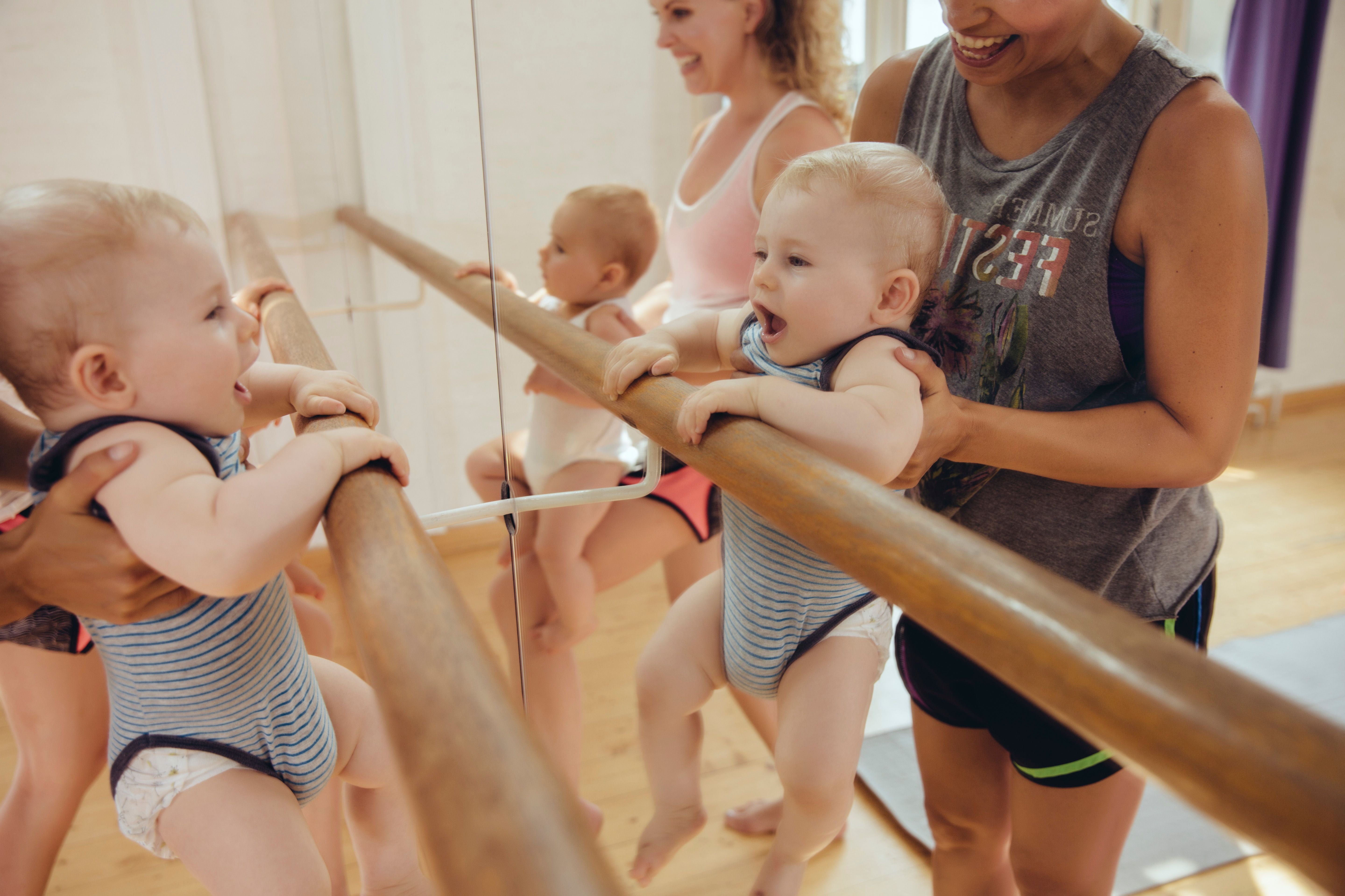 Zwei Babys in Stramplern halten sich an einer Ballettstange fest, begleitet von Erwachsenen.