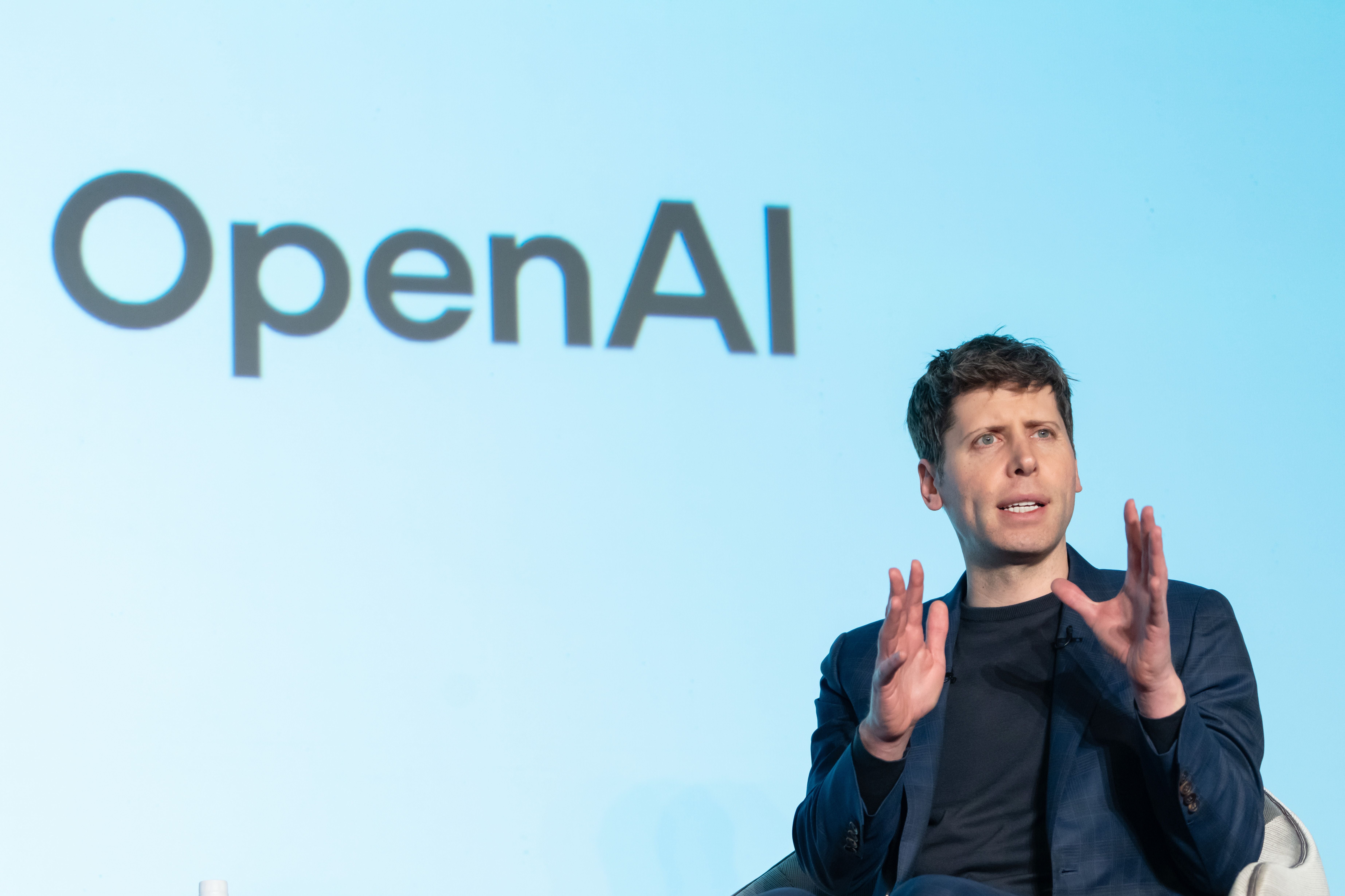 Ein Mann spricht auf einer Bühne vor einem großen OpenAI-Logo auf hellem Hintergrund.