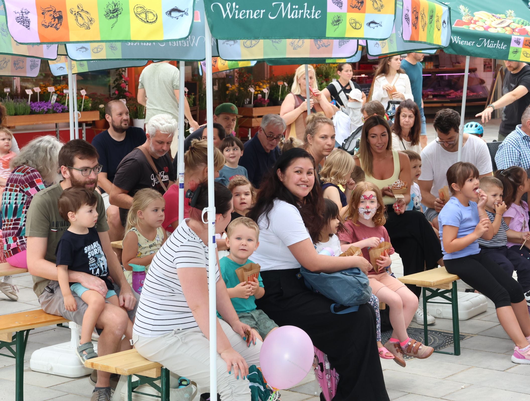 Gruppe von Erwachsenen und Kindern sitzt unter bunten Marktschirmen auf Bänken bei einem Markt.