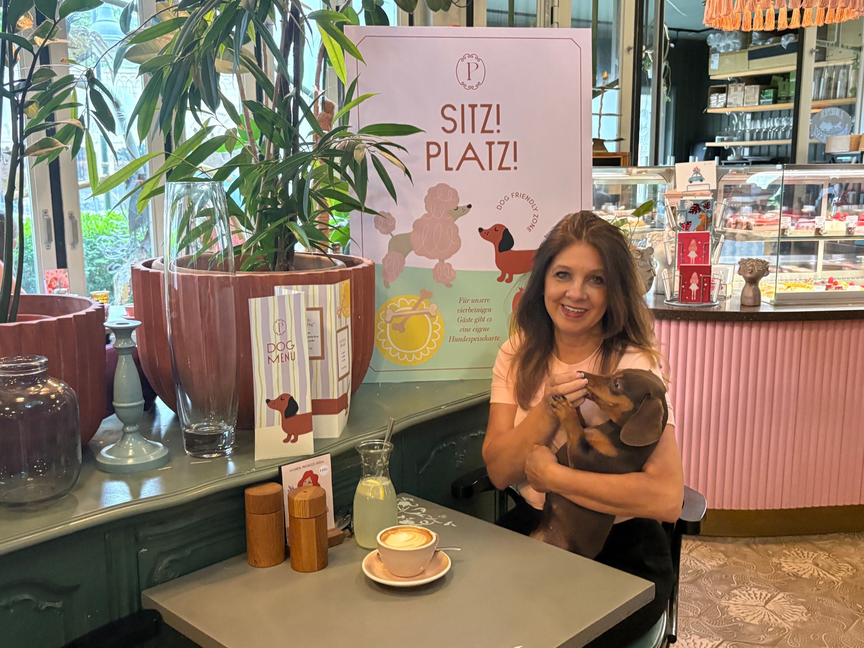 Frau sitzt mit einem Dackel auf dem Schoß in einem Café mit Hundemenü und Latte.