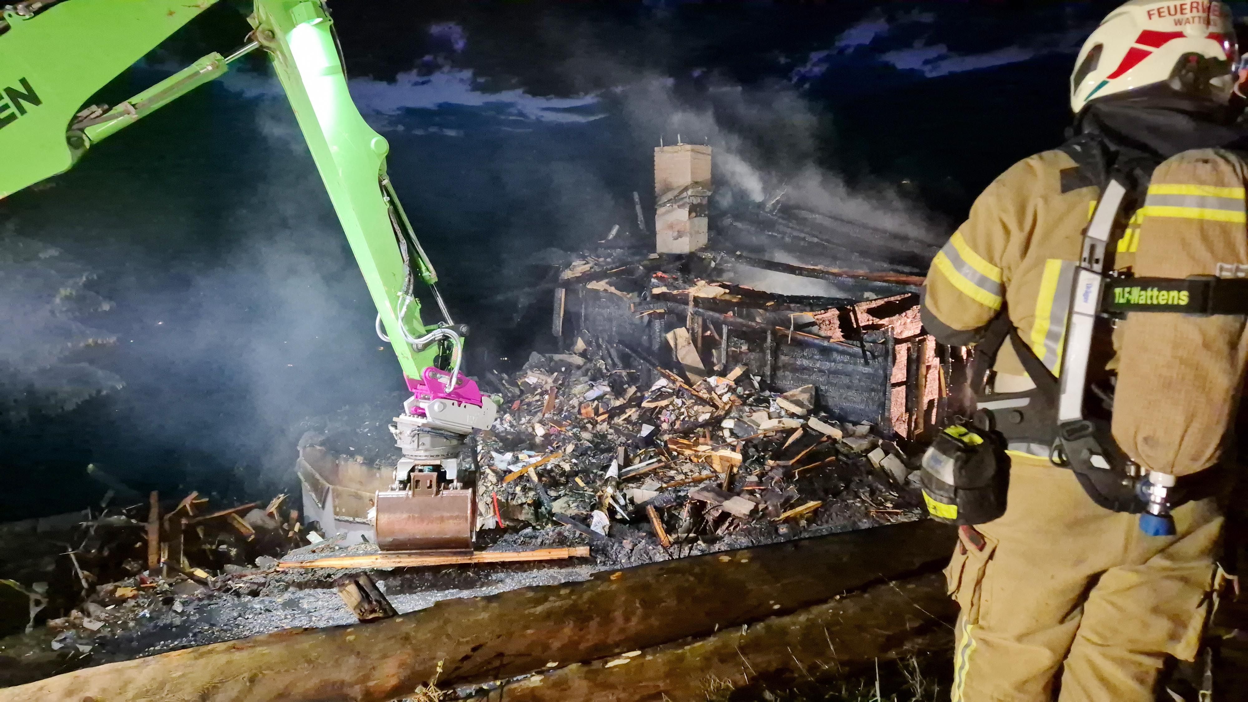Feuerwehr arbeitet nachts an den Trümmern eines abgebrannten Gebäudes mit Bagger und Ausrüstung.
