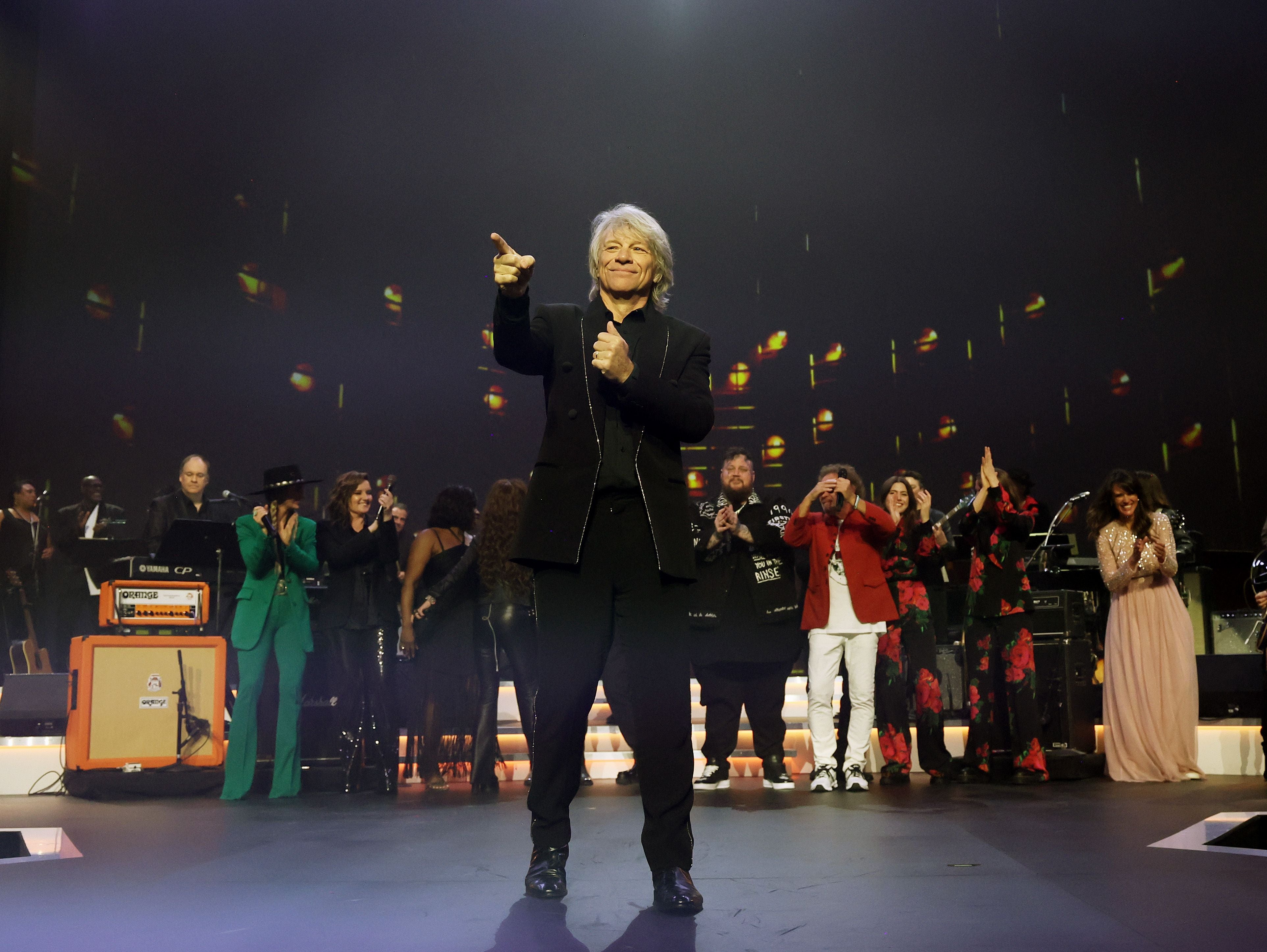 Jon Bon Jovi steht auf einer Bühne, applaudiert und wird von einer Gruppe applaudierender Menschen umgeben.