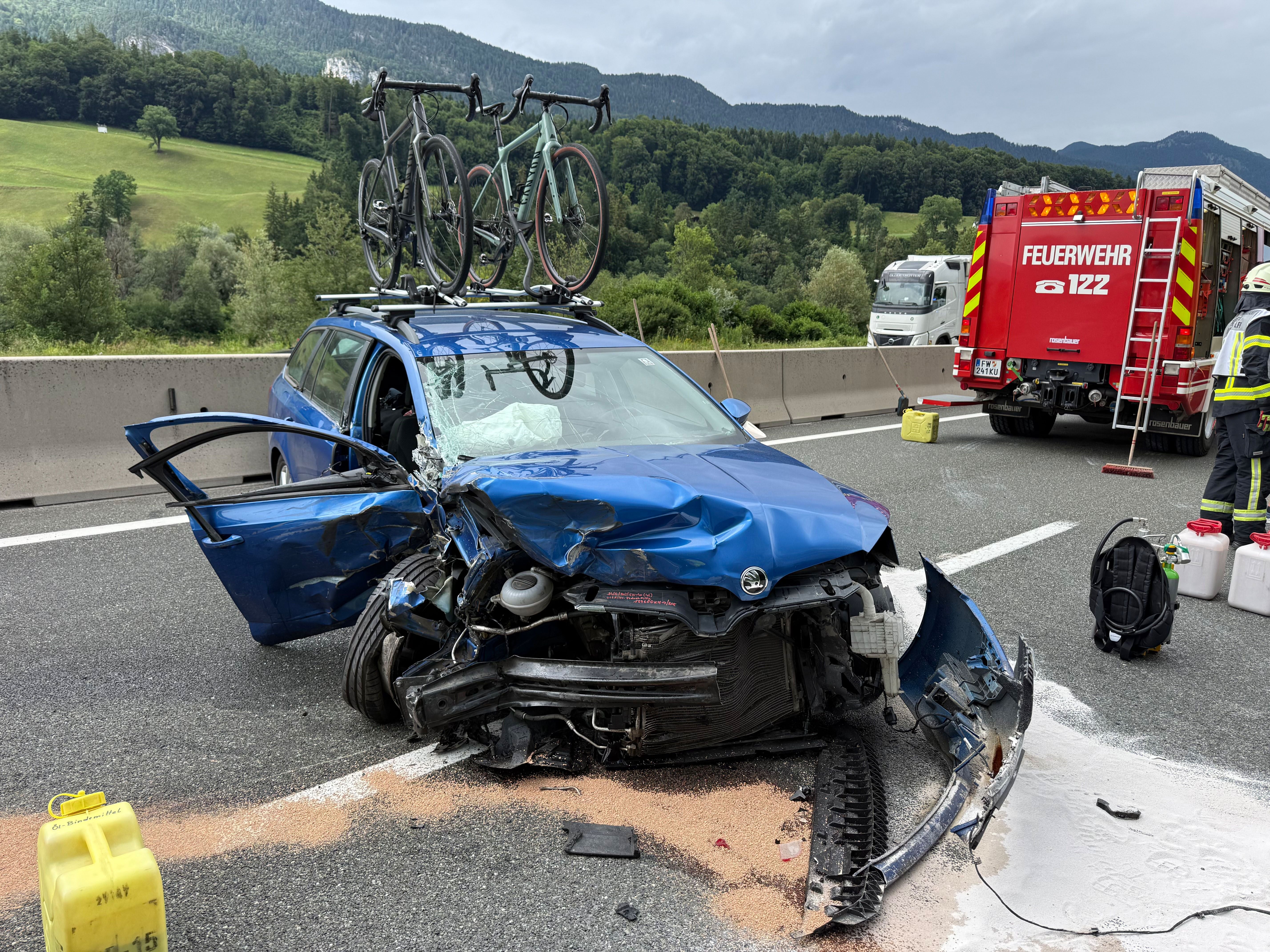 Schwer beschädigtes blaues Auto mit Fahrrädern auf dem Dach, Feuerwehr im Einsatz auf Autobahn.