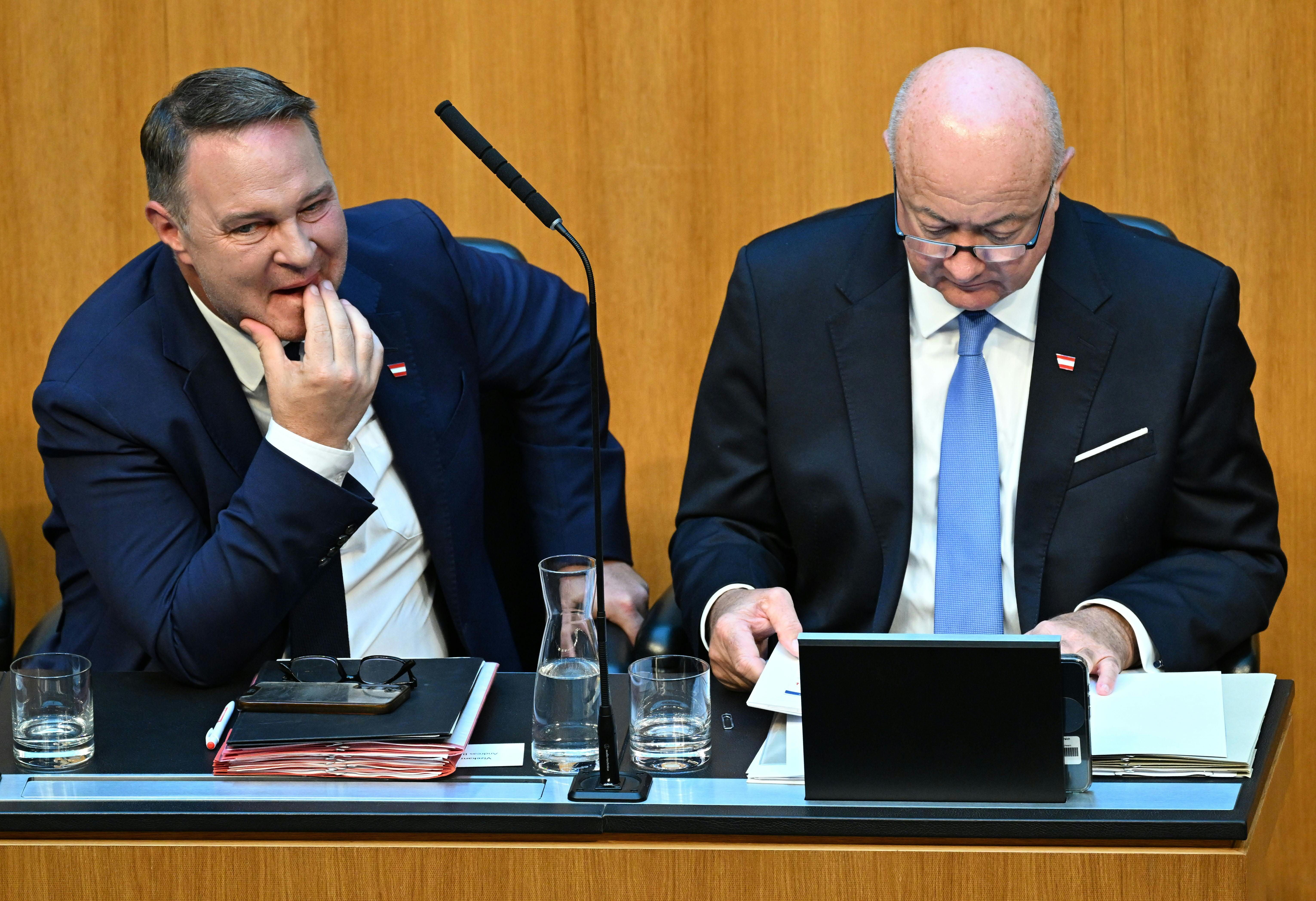 Zwei Männer im Anzug sitzen nebeneinander im Parlament und arbeiten an Unterlagen.