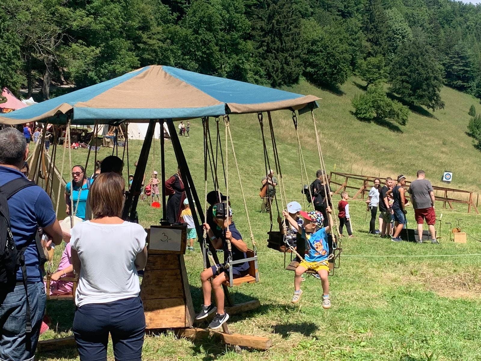 Kinder fahren auf einem Karussell beim Ritterfest auf einer Wiese mit vielen Besuchern im Hintergrund.