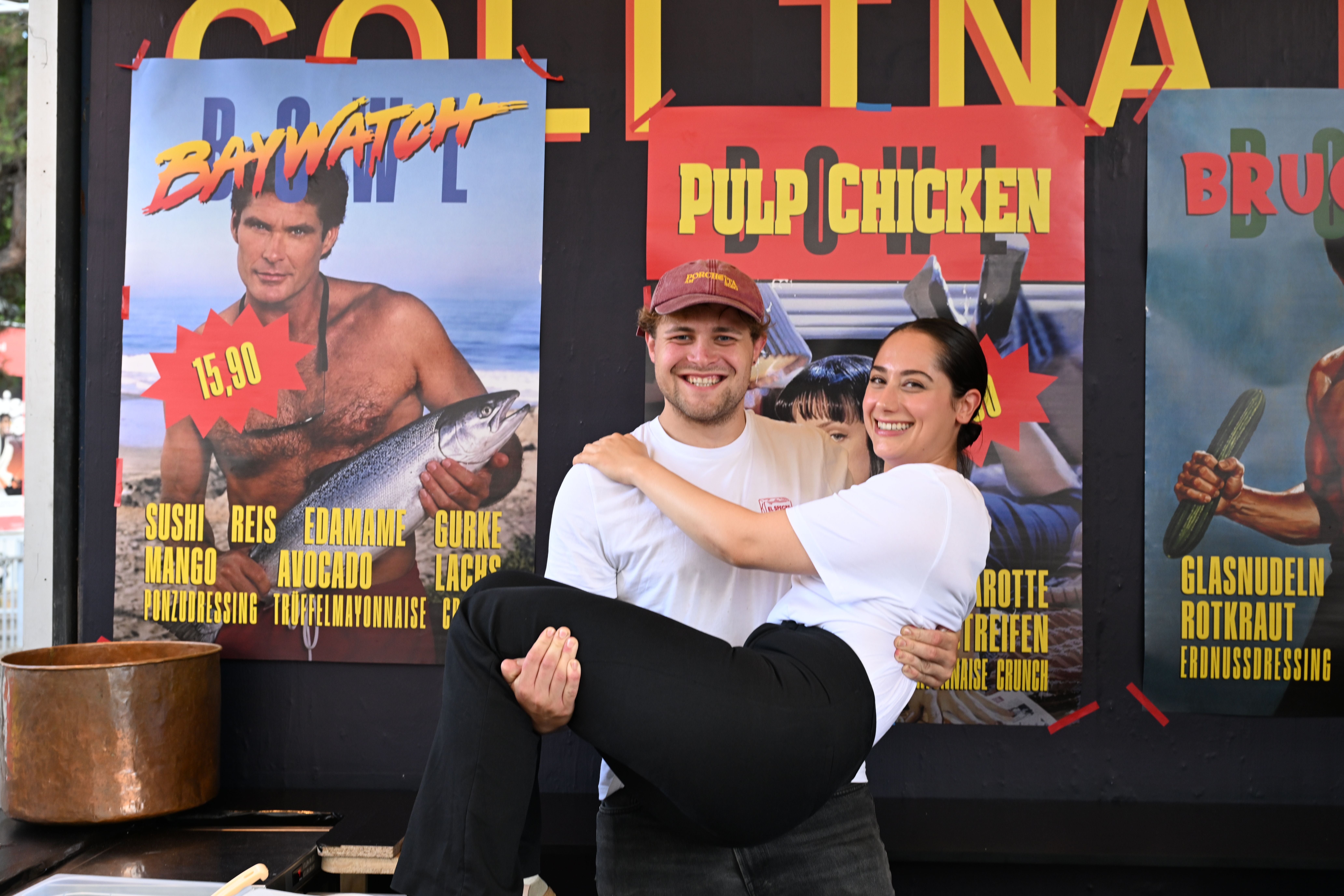 Ein Mann hält lachend eine Frau im Arm vor bunten Food-Postern auf einem Festival.