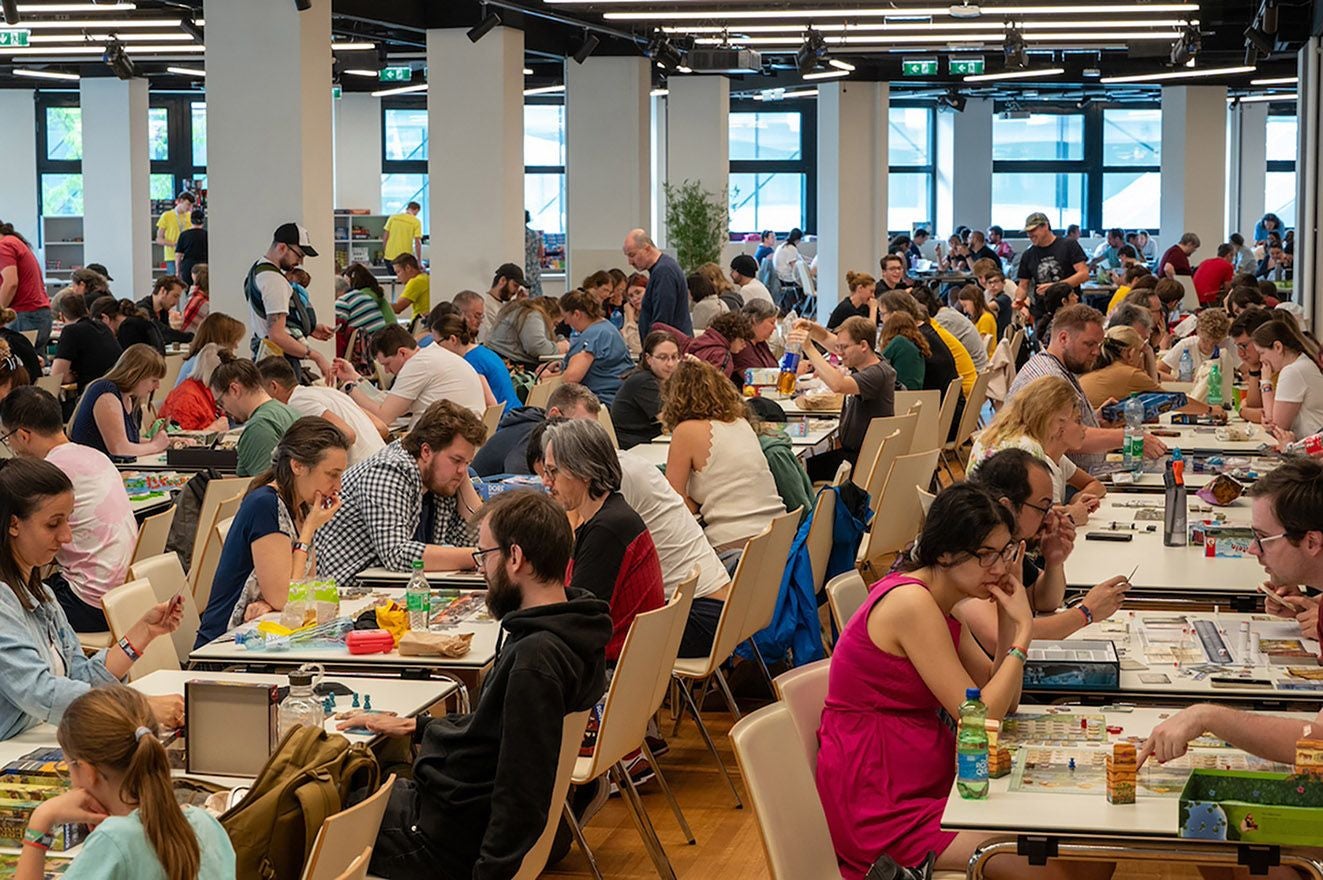 Viele Menschen sitzen an Tischen und spielen Brettspiele auf einem Spielefest in einer großen Halle.