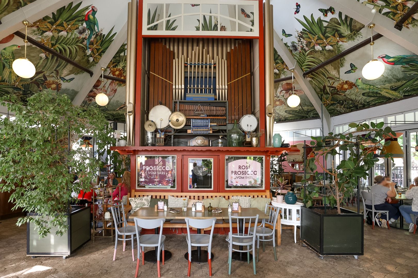 Bunt eingerichtetes Café mit Orgel, Pflanzen und verspieltem, tropischem Deckenmotiv.