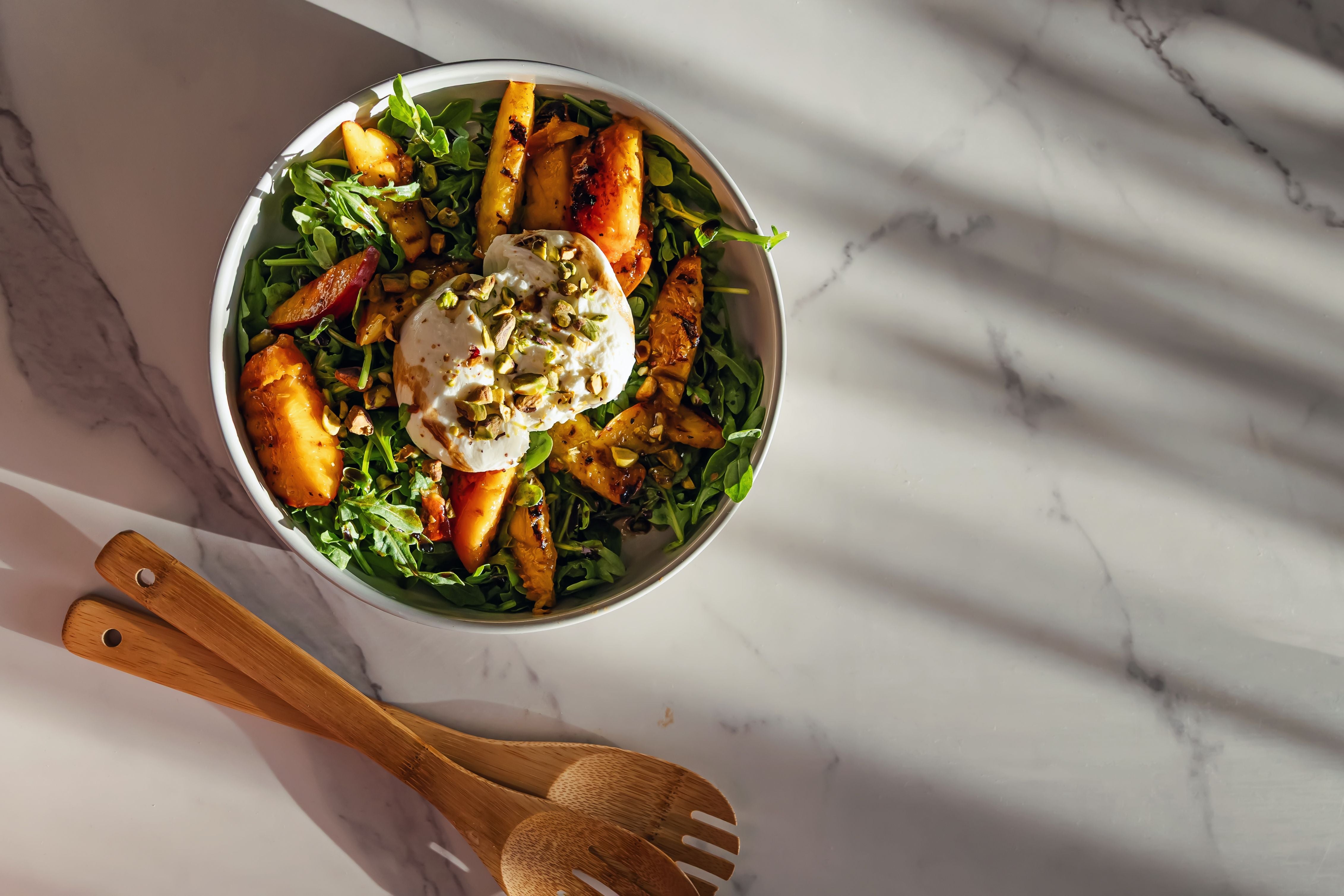 Salat mit gegrillten Pfirsichen, Burrata, Rucola und Pistazien in einer weißen Schüssel.