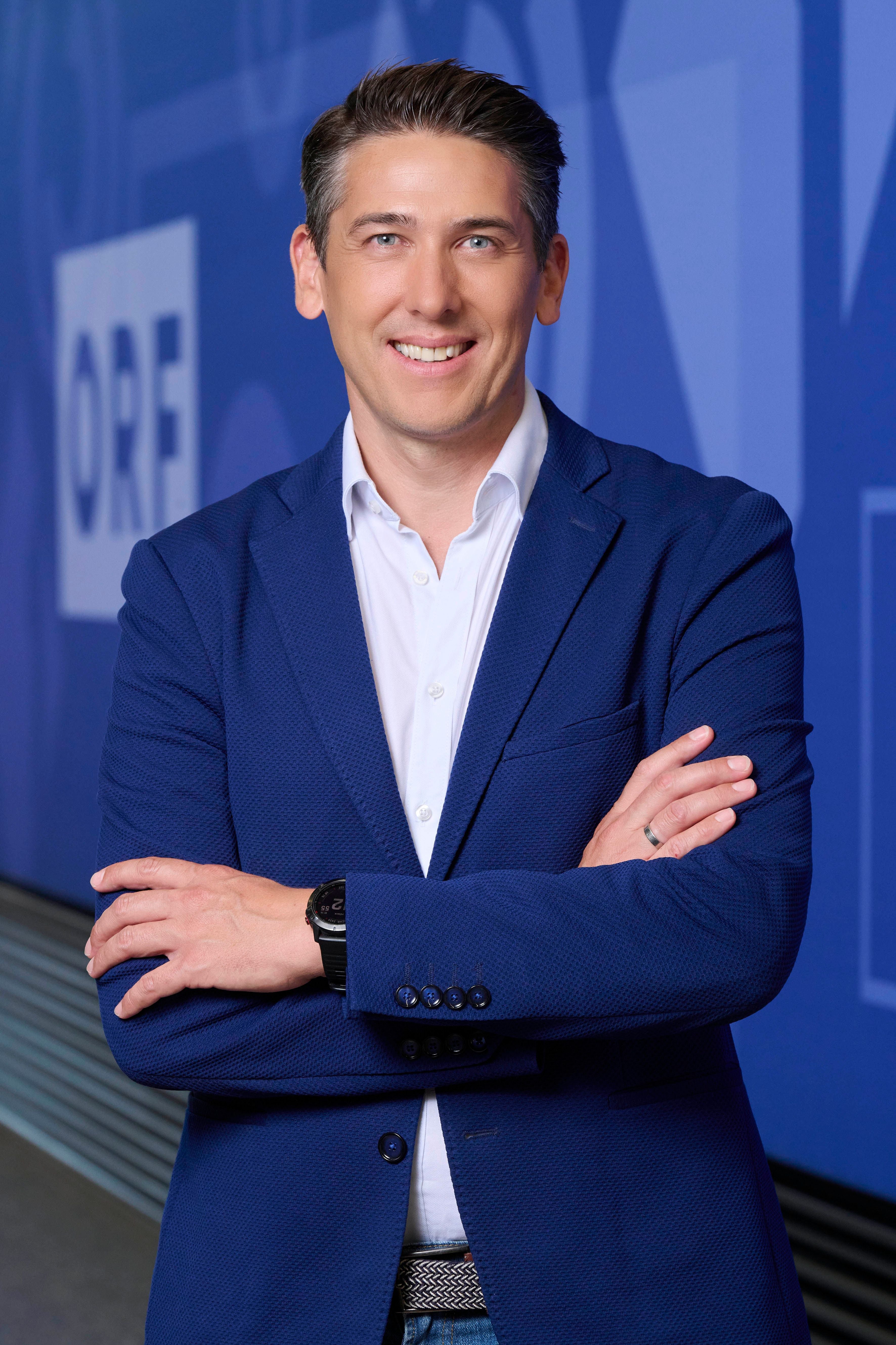 Mann im blauen Sakko lächelt mit verschränkten Armen vor blauem ORF-Hintergrund.