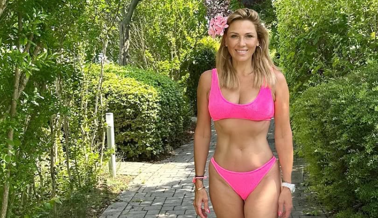 Frau im pinken Bikini steht auf einem Gartenweg, umgeben von grünen Büschen und Bäumen.