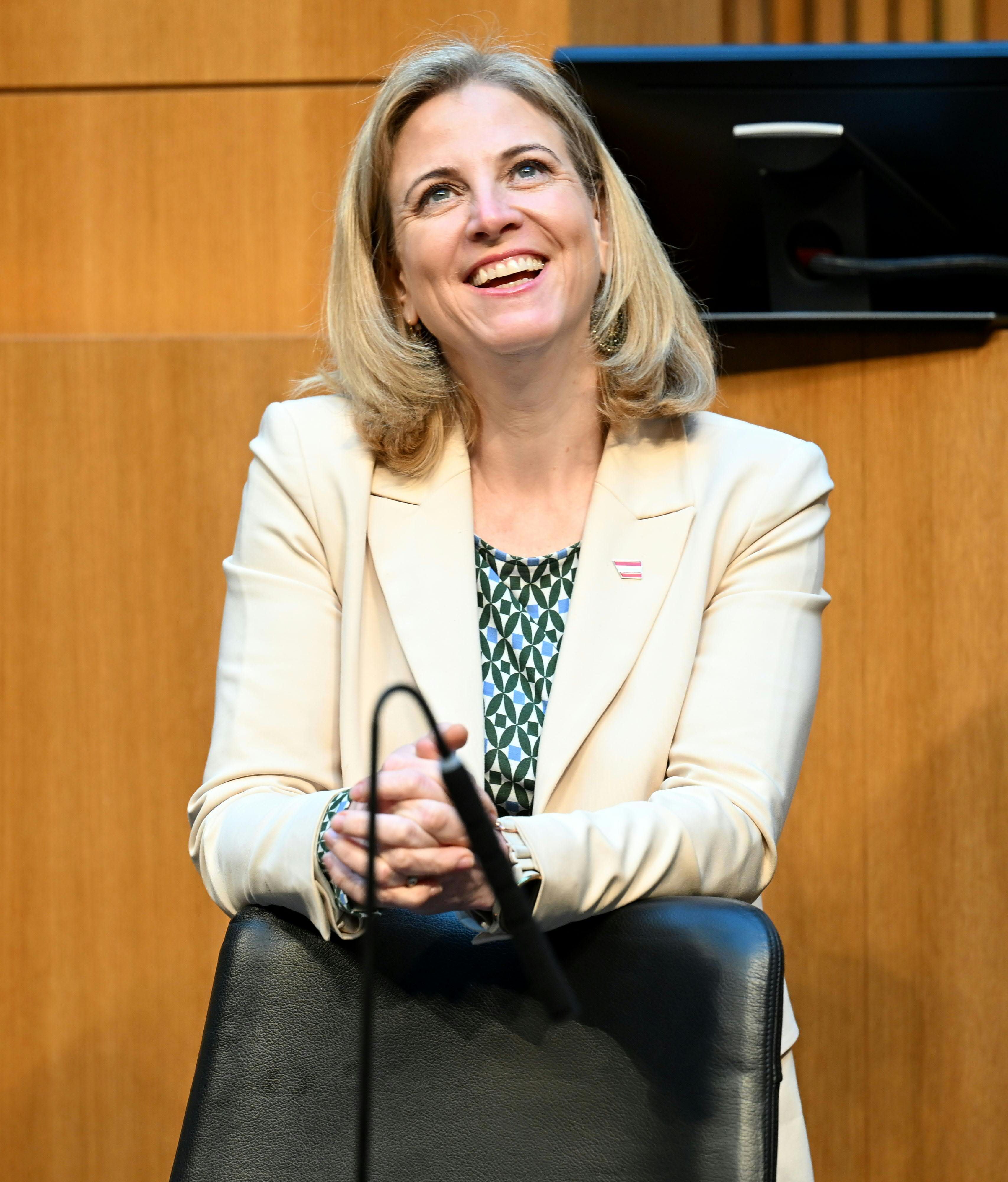 Frau mit blondem Haar und hellem Blazer lächelt im Parlament vor Holzverkleidung.