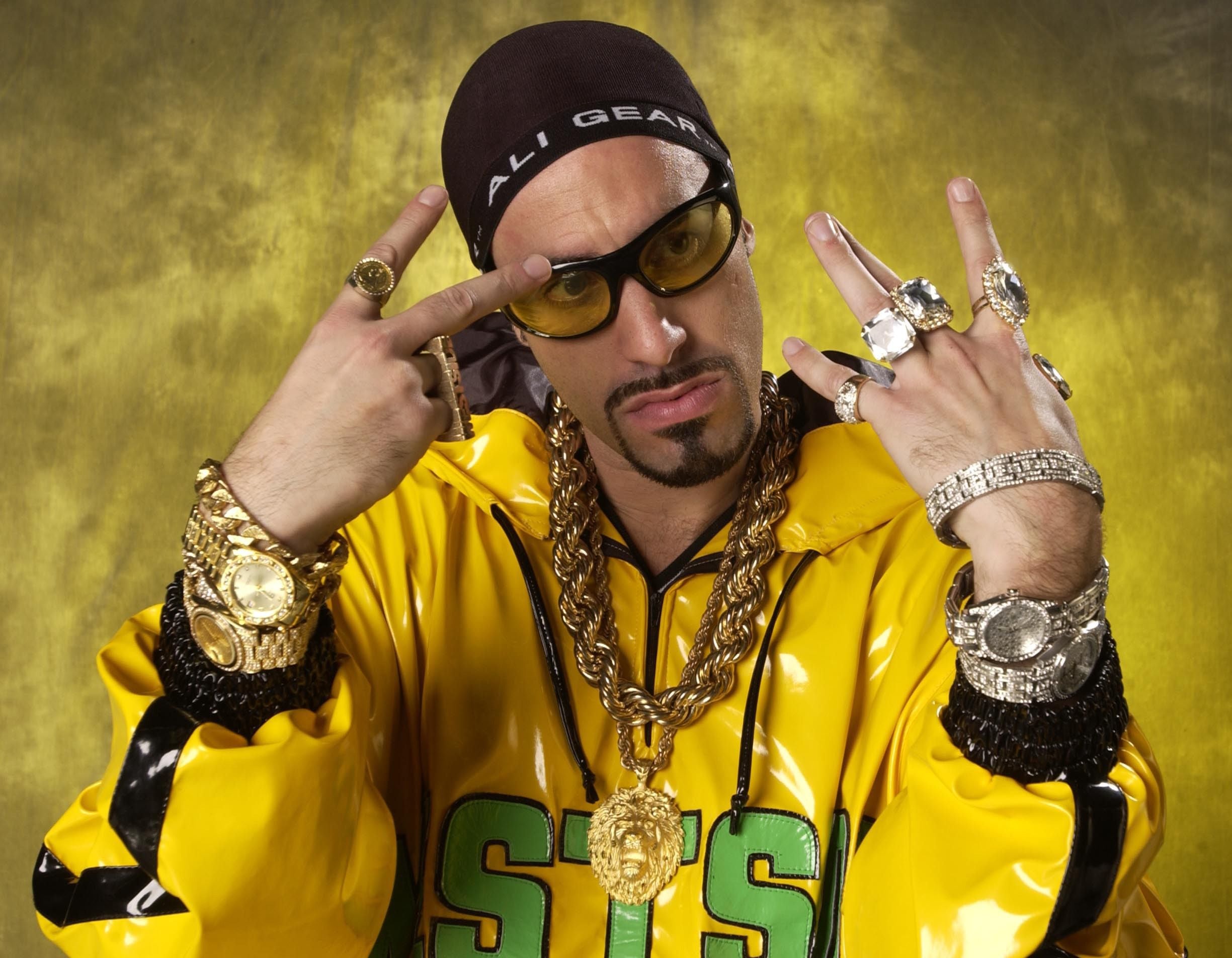 Mann mit gelber Jacke, viel Goldschmuck und Sonnenbrille posiert vor gelbem Hintergrund.