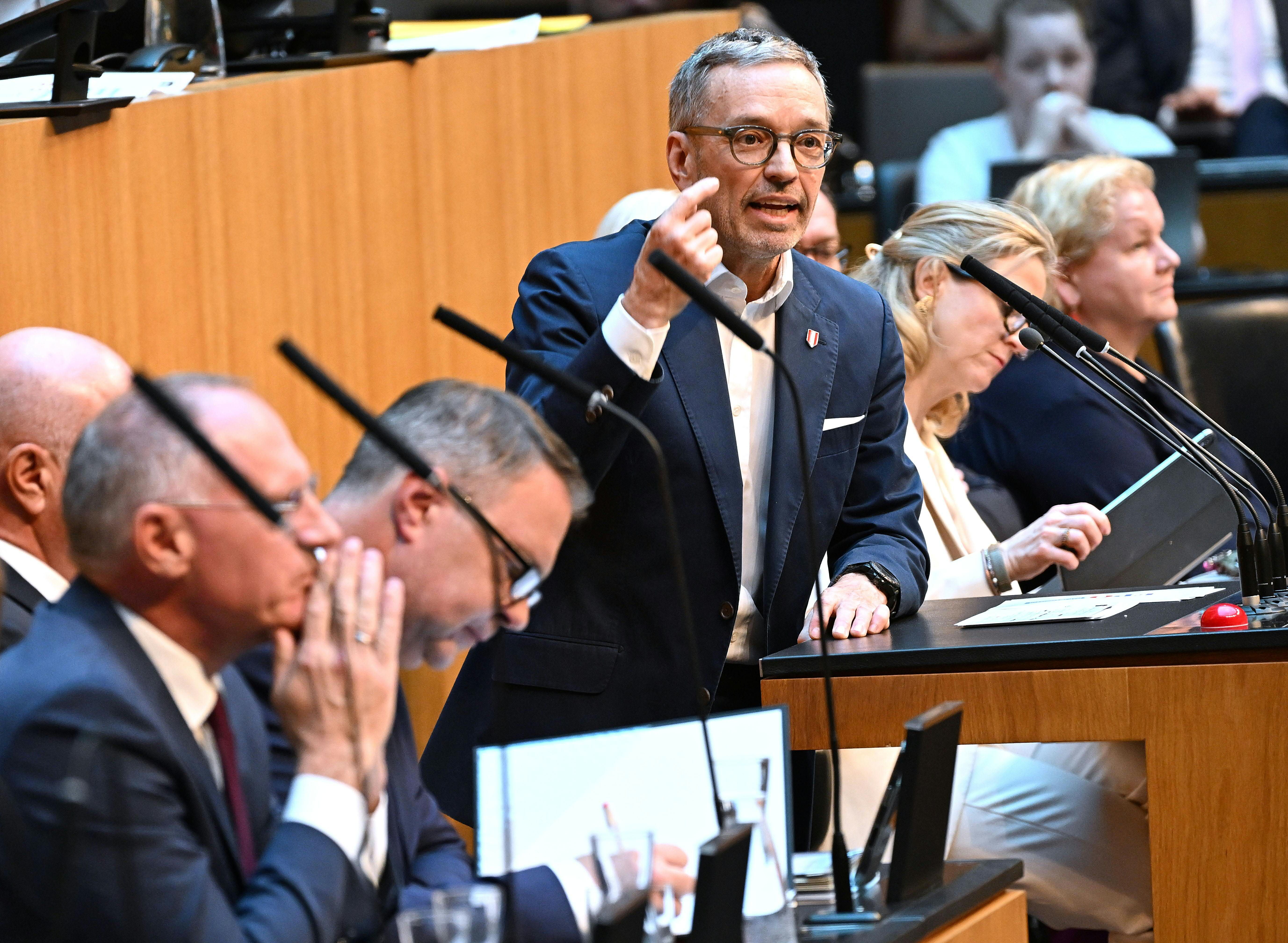 Ein Mann im Anzug spricht engagiert bei einer Parlamentssitzung in einem modernen Saal.