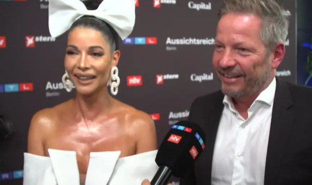 Eine Frau mit großem weißen Haarband und ein Mann werden auf dem roten Teppich interviewt.