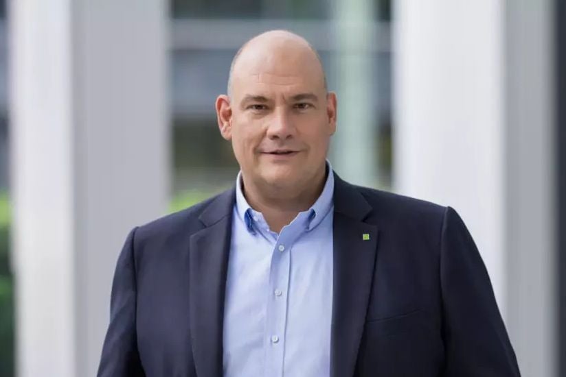 Mann im Anzug mit Glatze steht vor einem modernen, hellen Hintergrund.