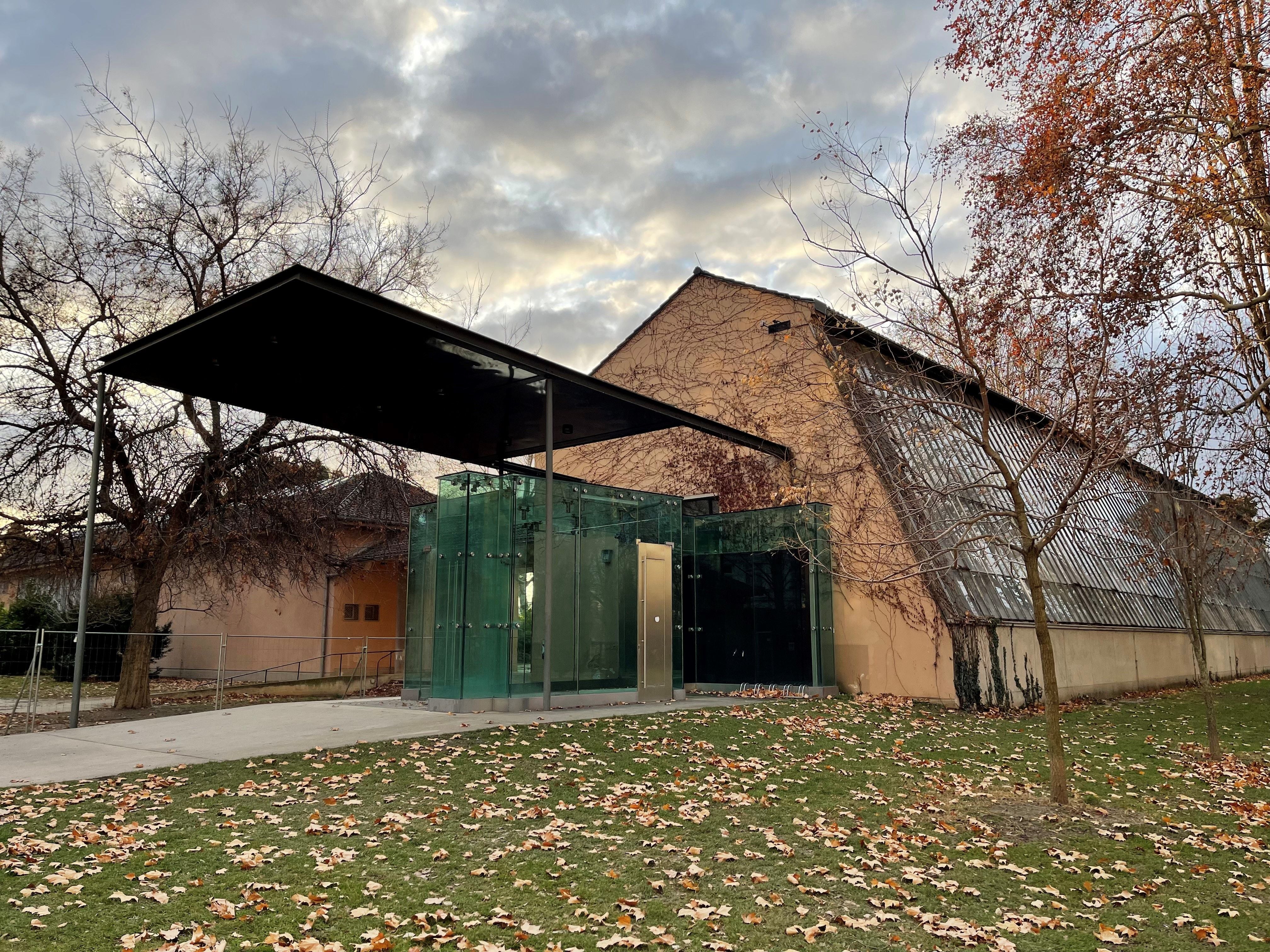 Modernes Gebäude mit Glasfassade und Pavillon, umgeben von herbstlichen Bäumen und fallenden Blättern.