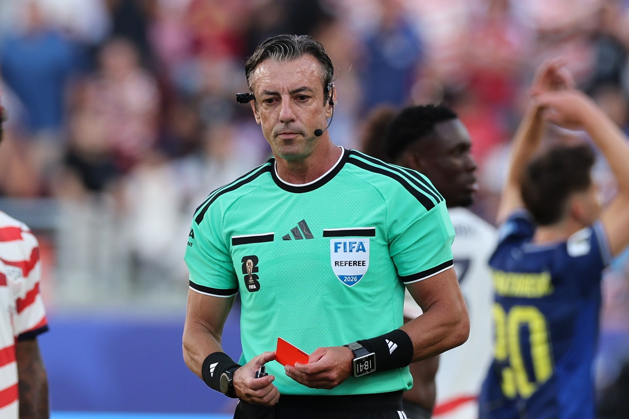 Ein Fußballschiedsrichter zeigt eine rote Karte während eines Spiels, Menschen sind im Hintergrund.
