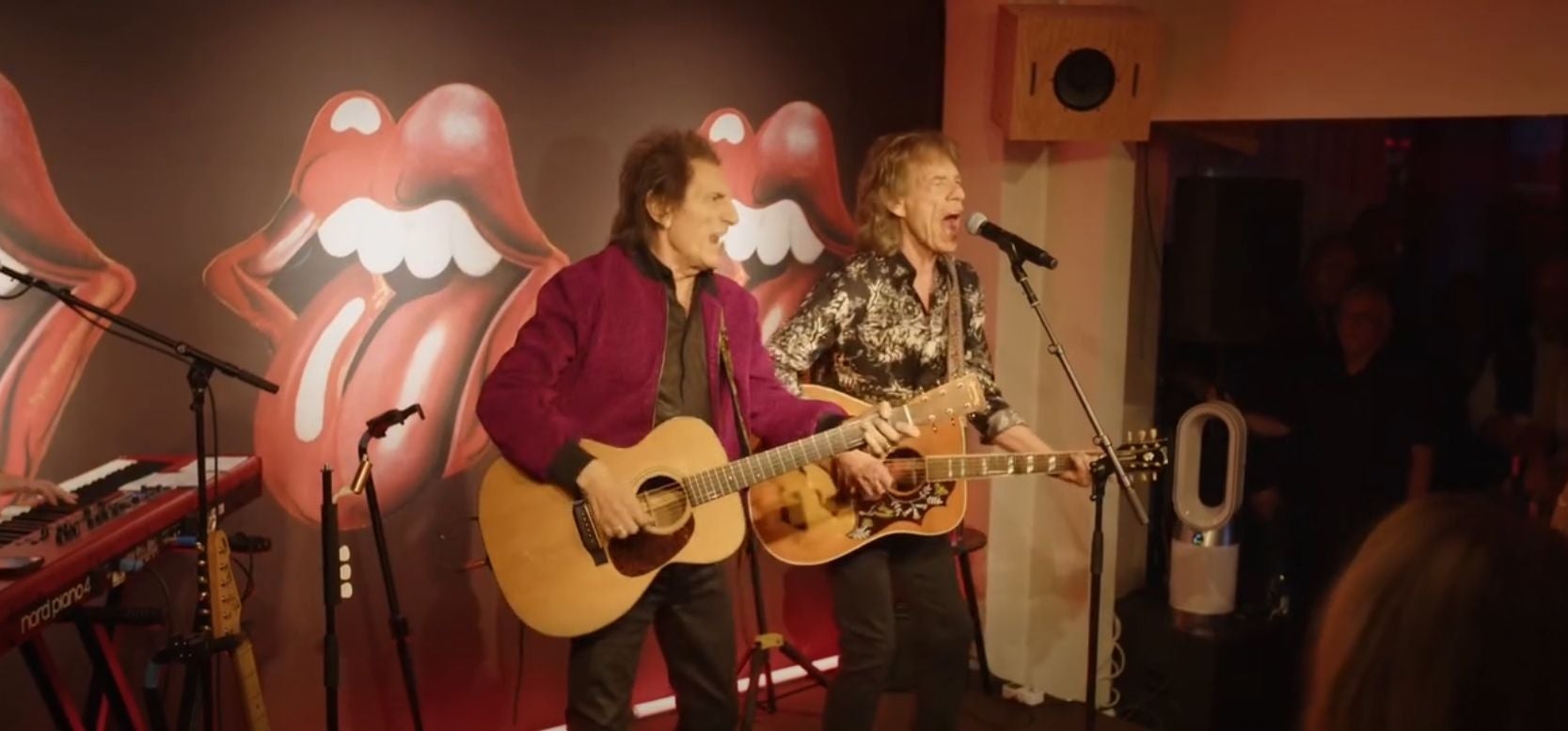 Zwei Musiker der Rolling Stones spielen Gitarre auf einer Bühne mit Zungenlogo im Hintergrund.