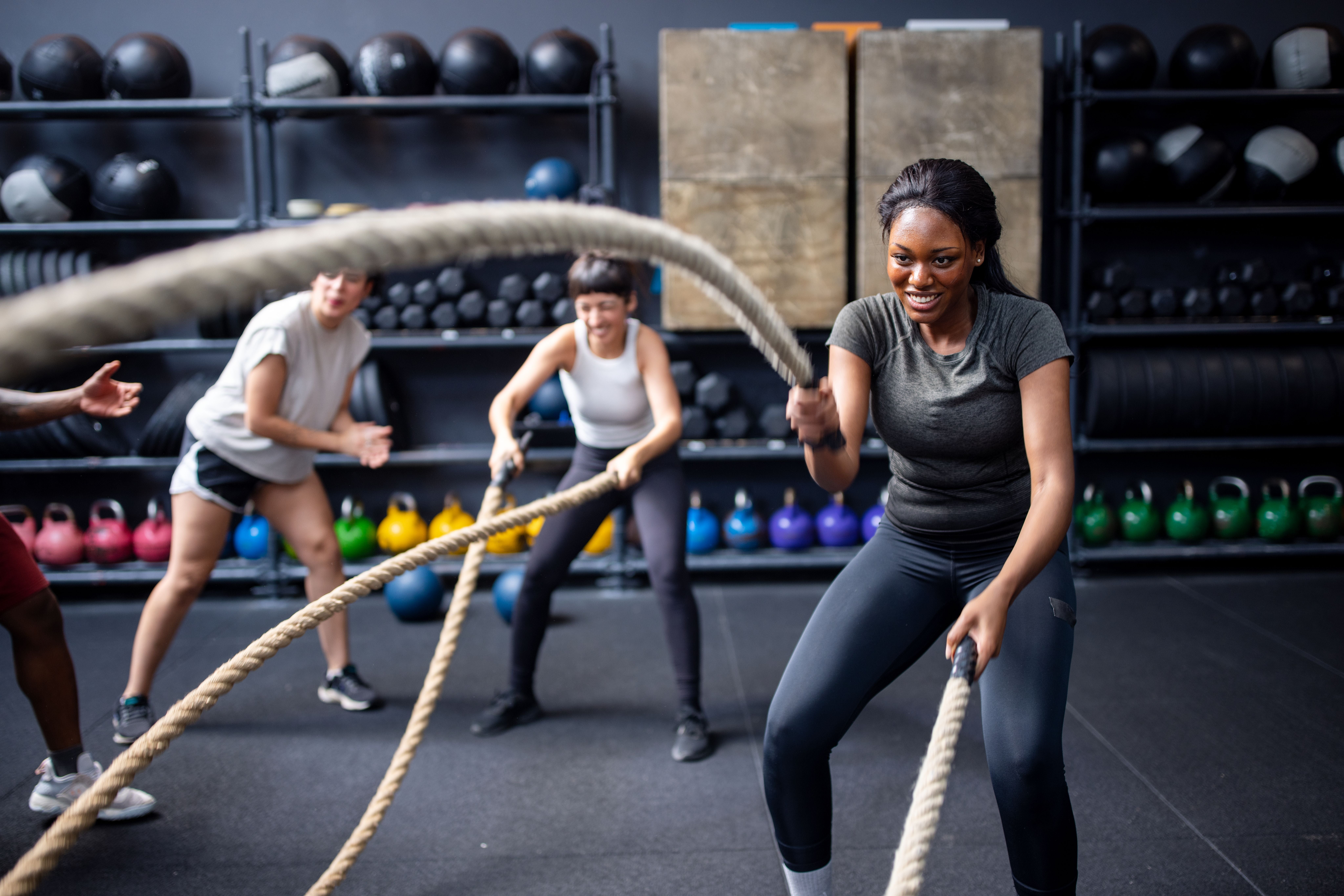 Drei Frauen trainieren mit Battle Ropes in einem Fitnessstudio, Freunde feuern sie an.