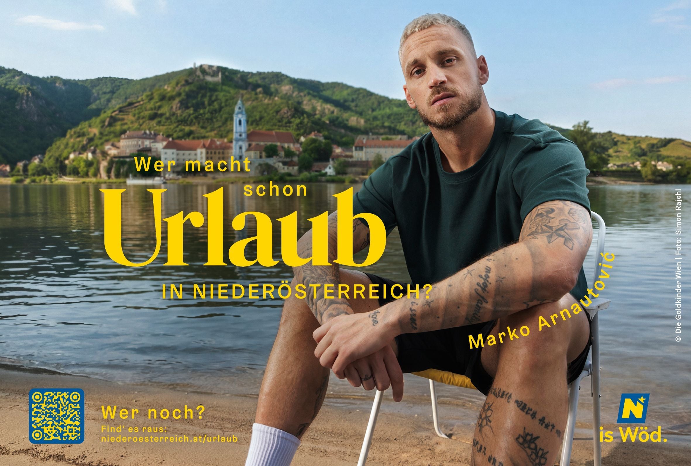 Ein tätowierter Mann sitzt am Seeufer vor einer malerischen Kulisse mit Bergen und Kirchen.