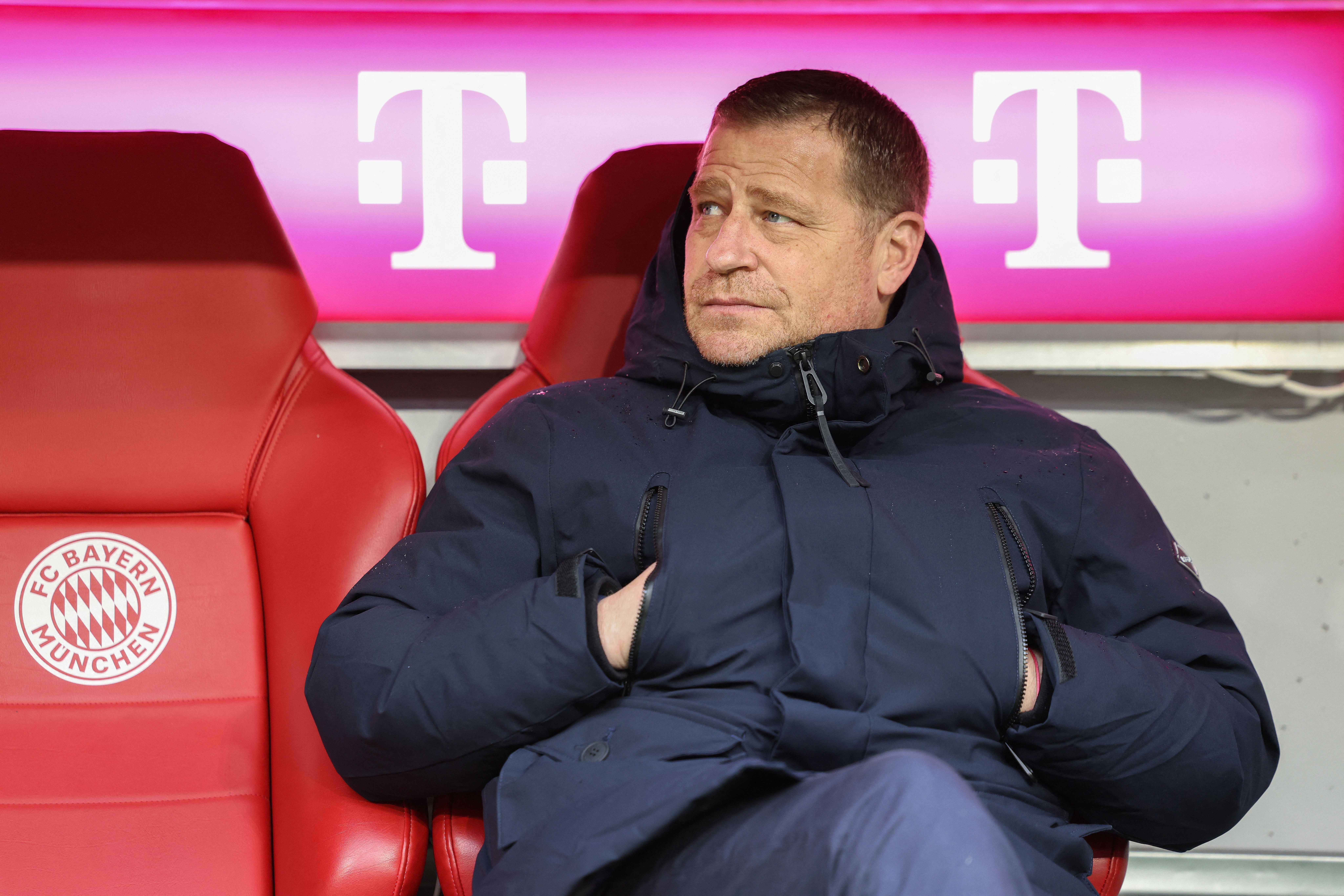 Ein Mann sitzt mit dunkler Jacke auf der FC Bayern München Bank vor pinkem Hintergrund.