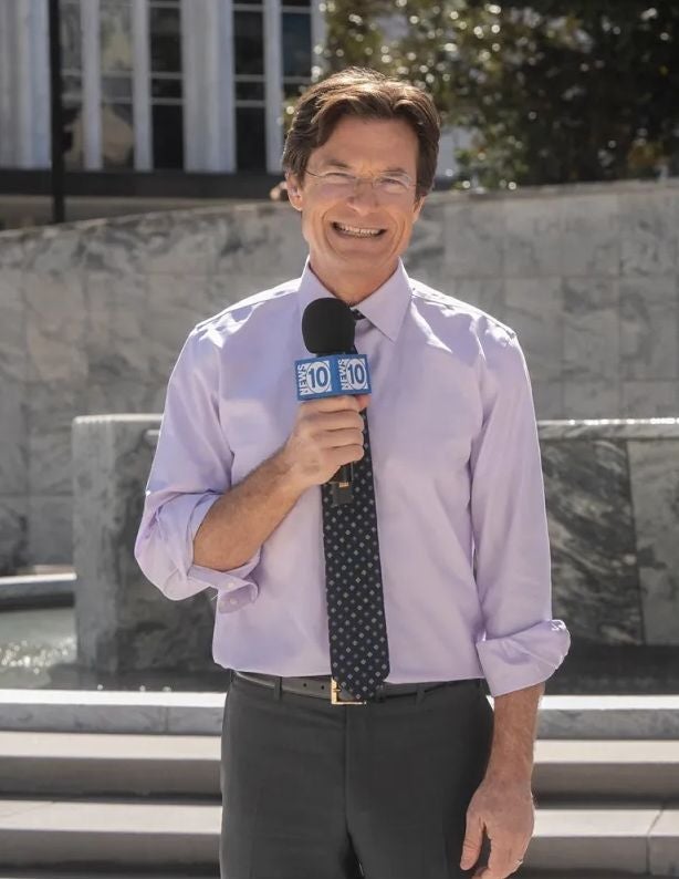 Ein lächelnder Reporter mit Mikrofon steht im Freien vor einem steinernen Brunnen.