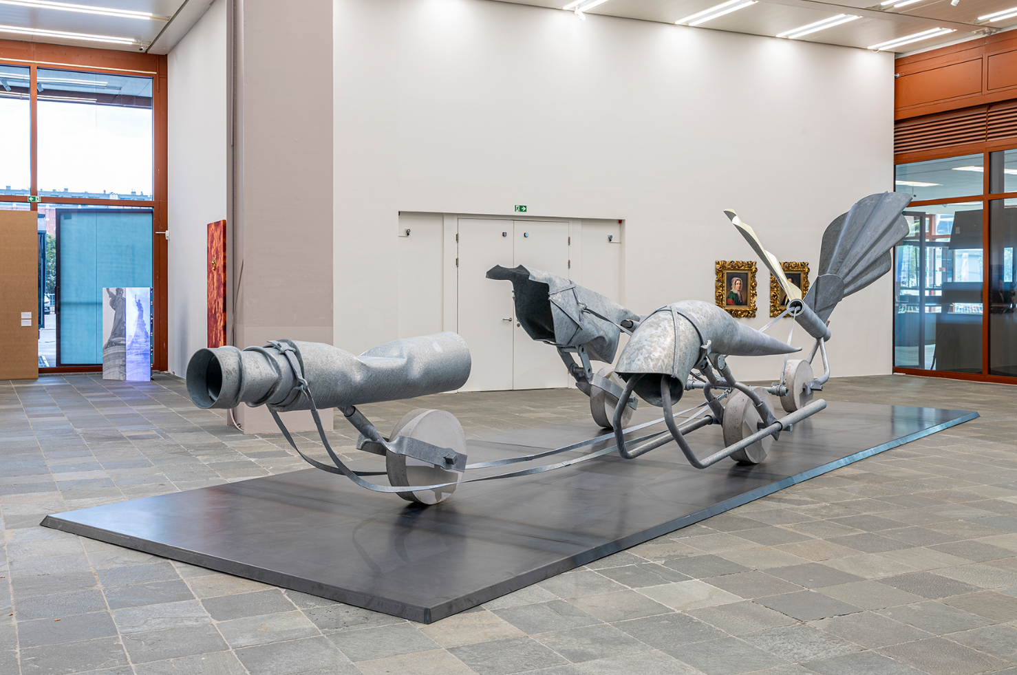 Große abstrakte Skulptur aus Metallteilen steht auf einer Plattform in einem modernen Ausstellungsraum.