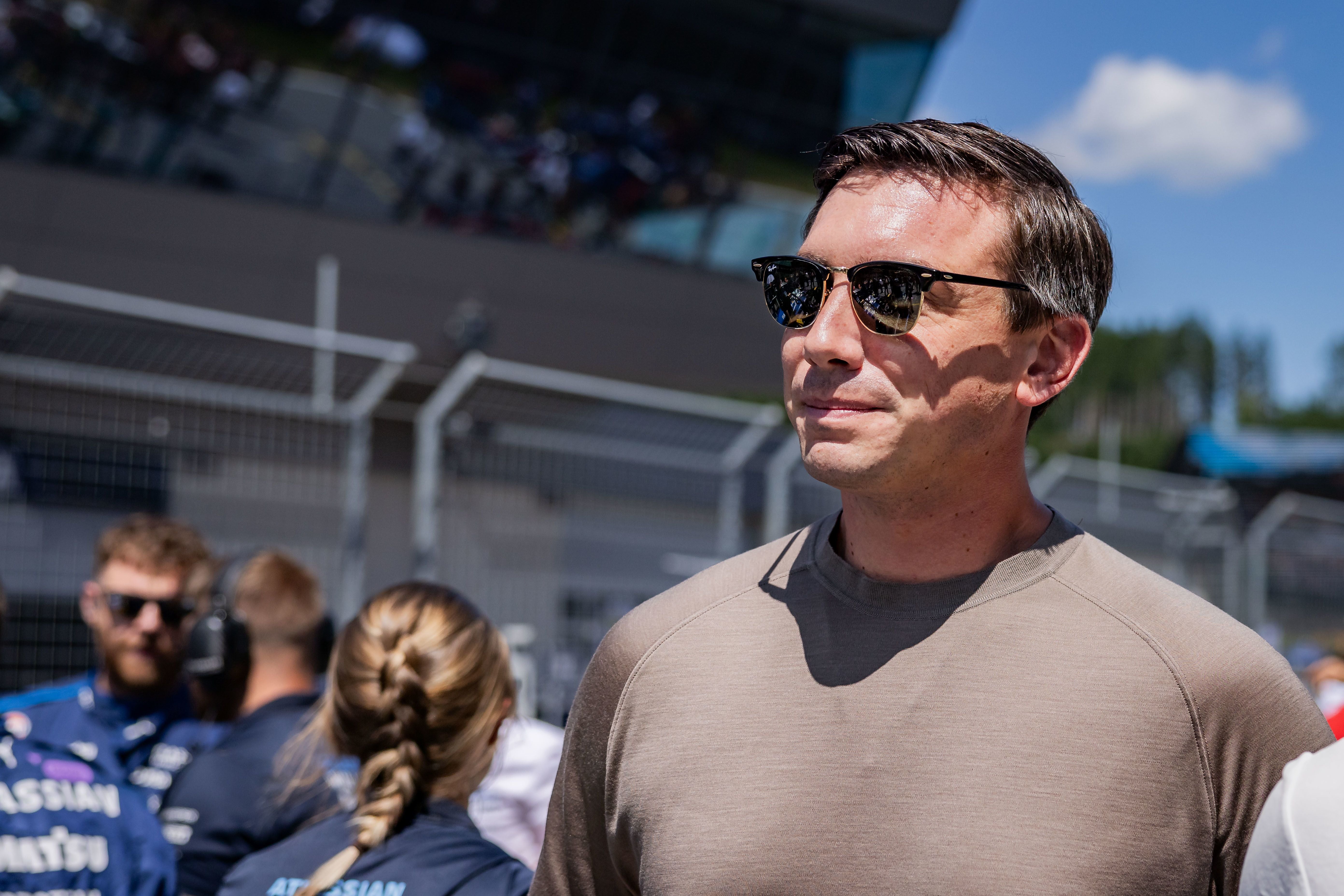 Ein Mann mit Sonnenbrille steht lächelnd im Fahrerlager einer Formel-1-Rennstrecke.