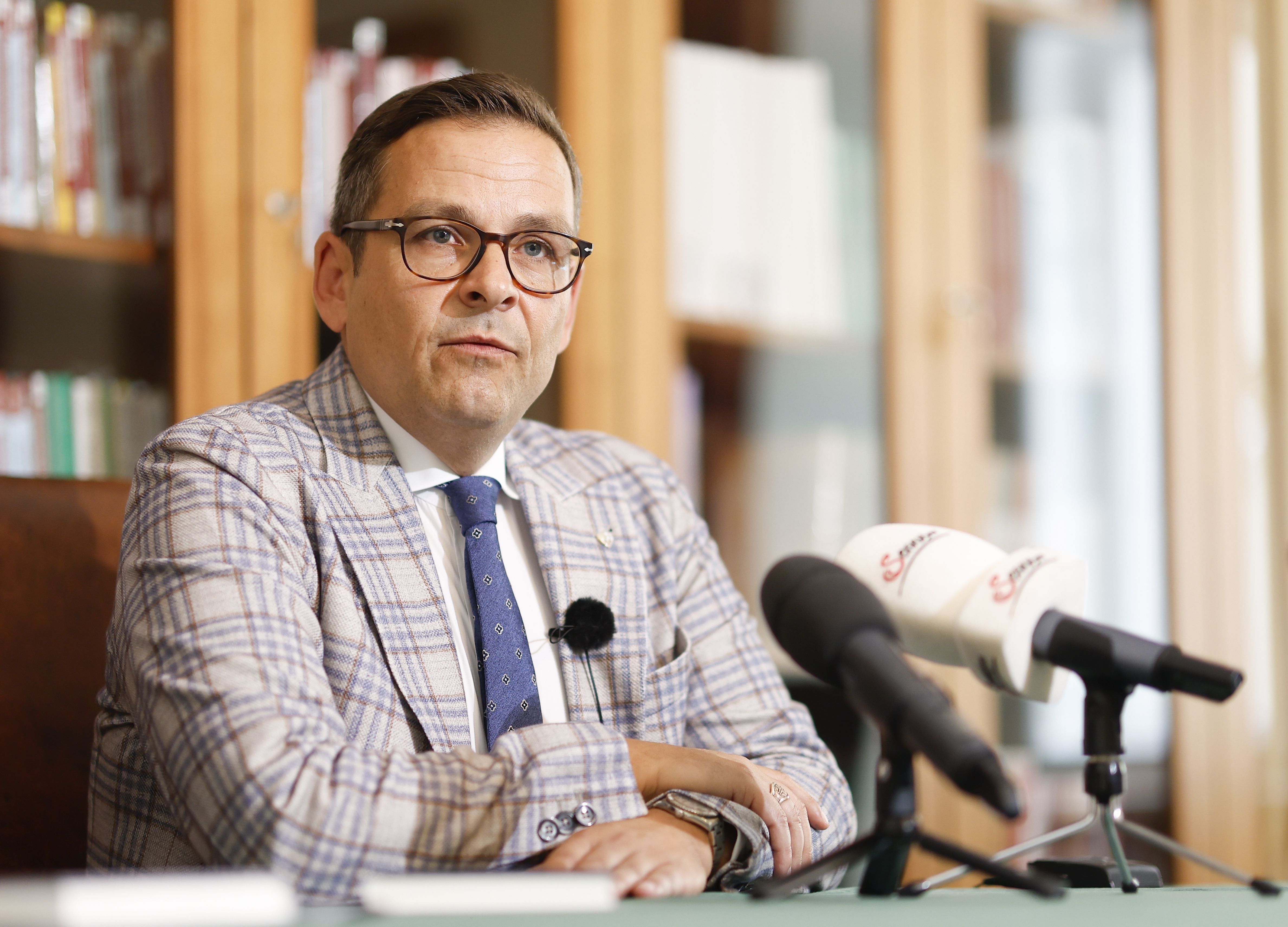 Ein Mann im karierten Anzug spricht bei einer Pressekonferenz vor Mikrofonen.