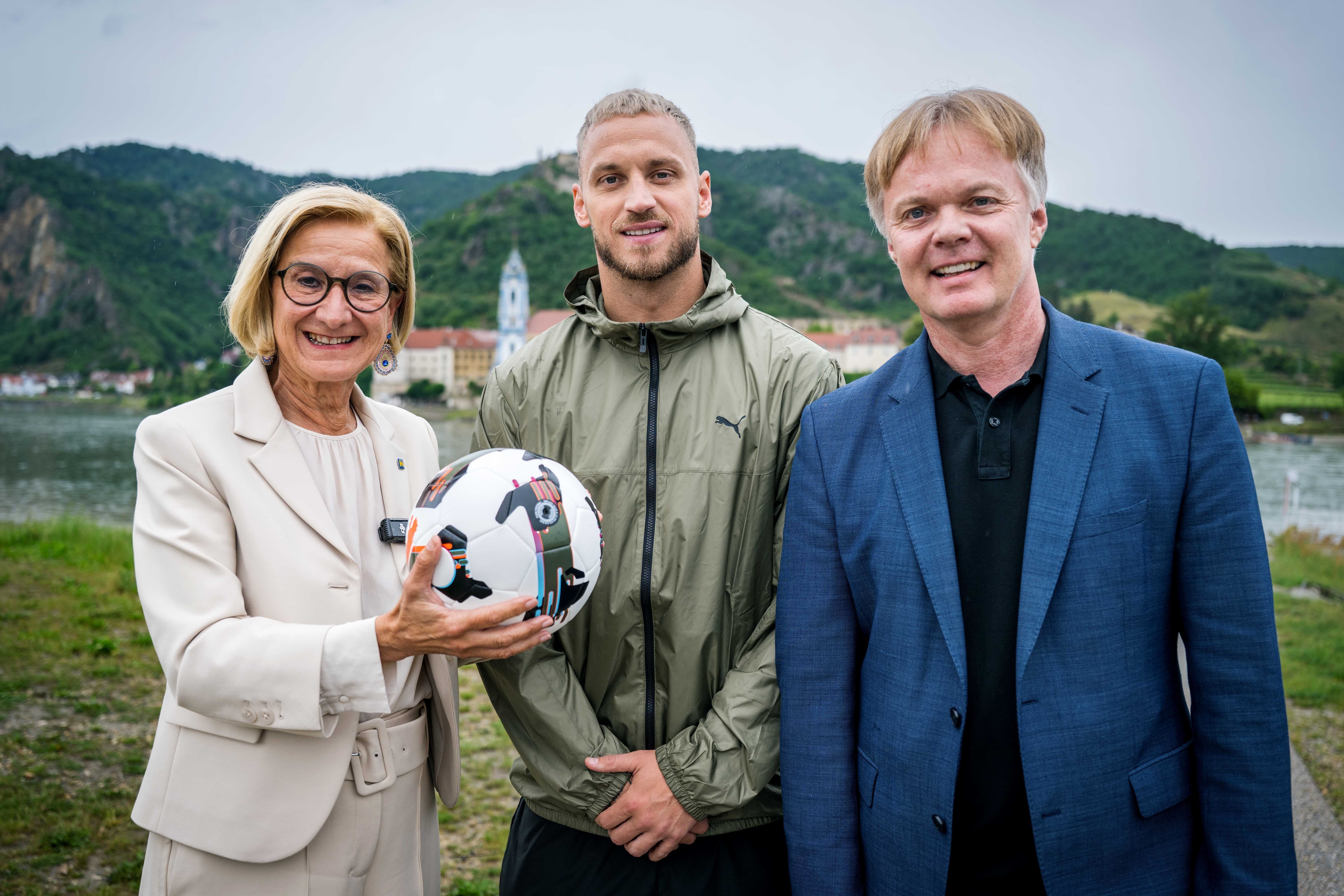 Drei Personen posieren draußen, eine Frau hält einen Fußball in der Hand.
