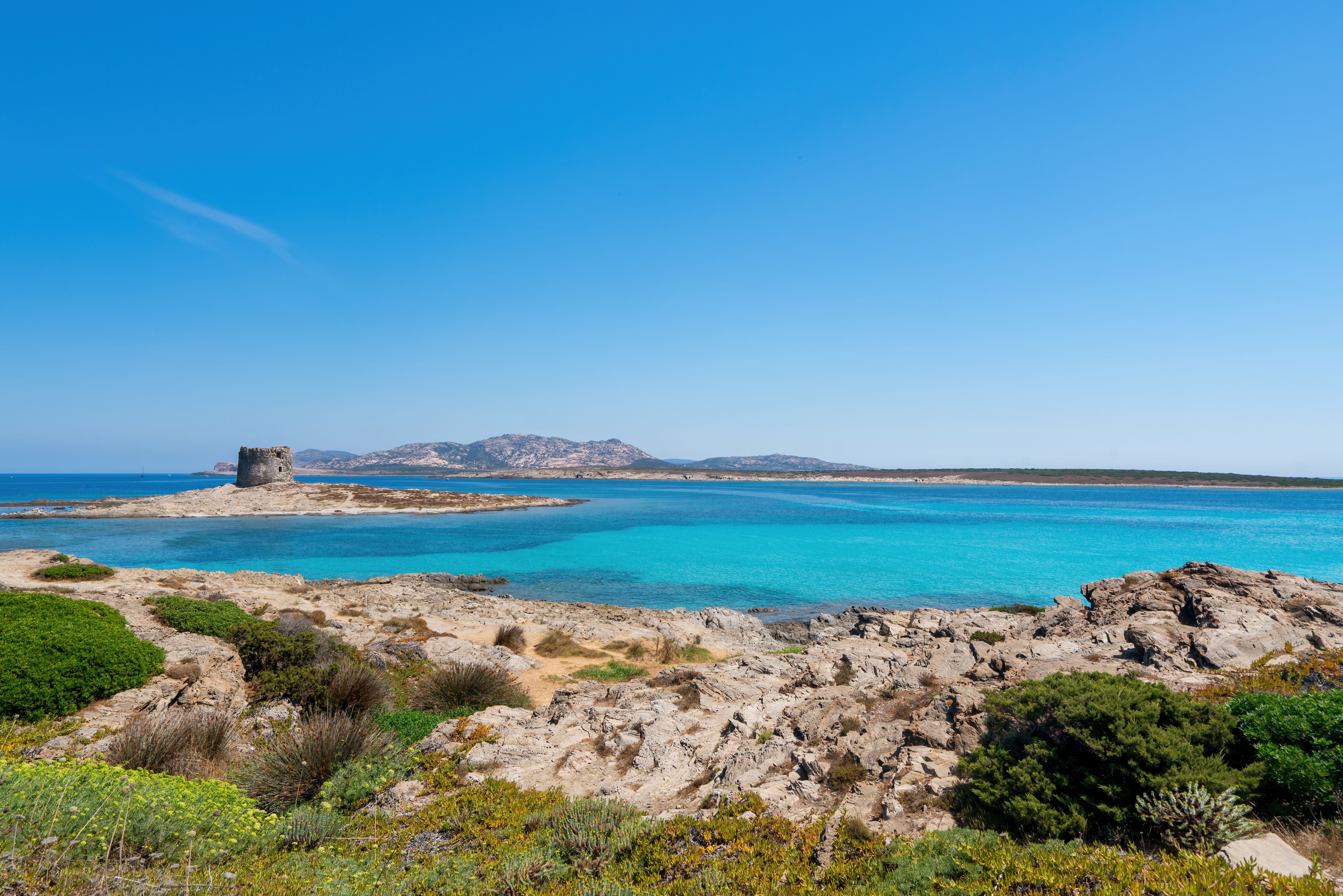 Felsige Küste mit türkisblauem Meer und historischem Turm am Strand La Pelosa, Sardinien.