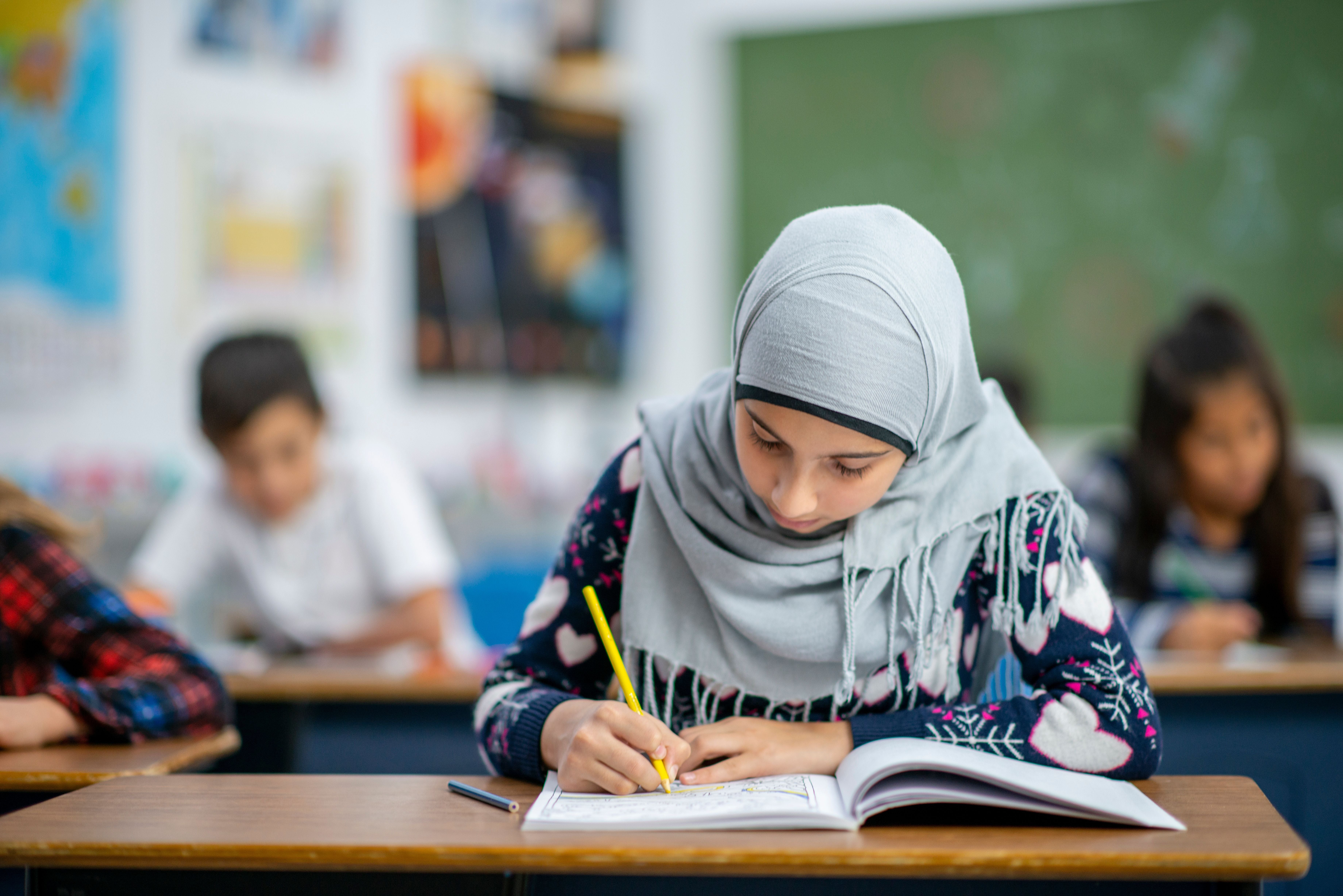 Ein Mädchen mit Kopftuch sitzt konzentriert an ihrem Schultisch und schreibt in ein Heft. Sie trägt einen dunkelblauen Pullover mit Herzmustern. Im Hintergrund sitzen weitere Schulkinder in einem Klassenzimmer mit Tafel und bunten Postern an der Wand.