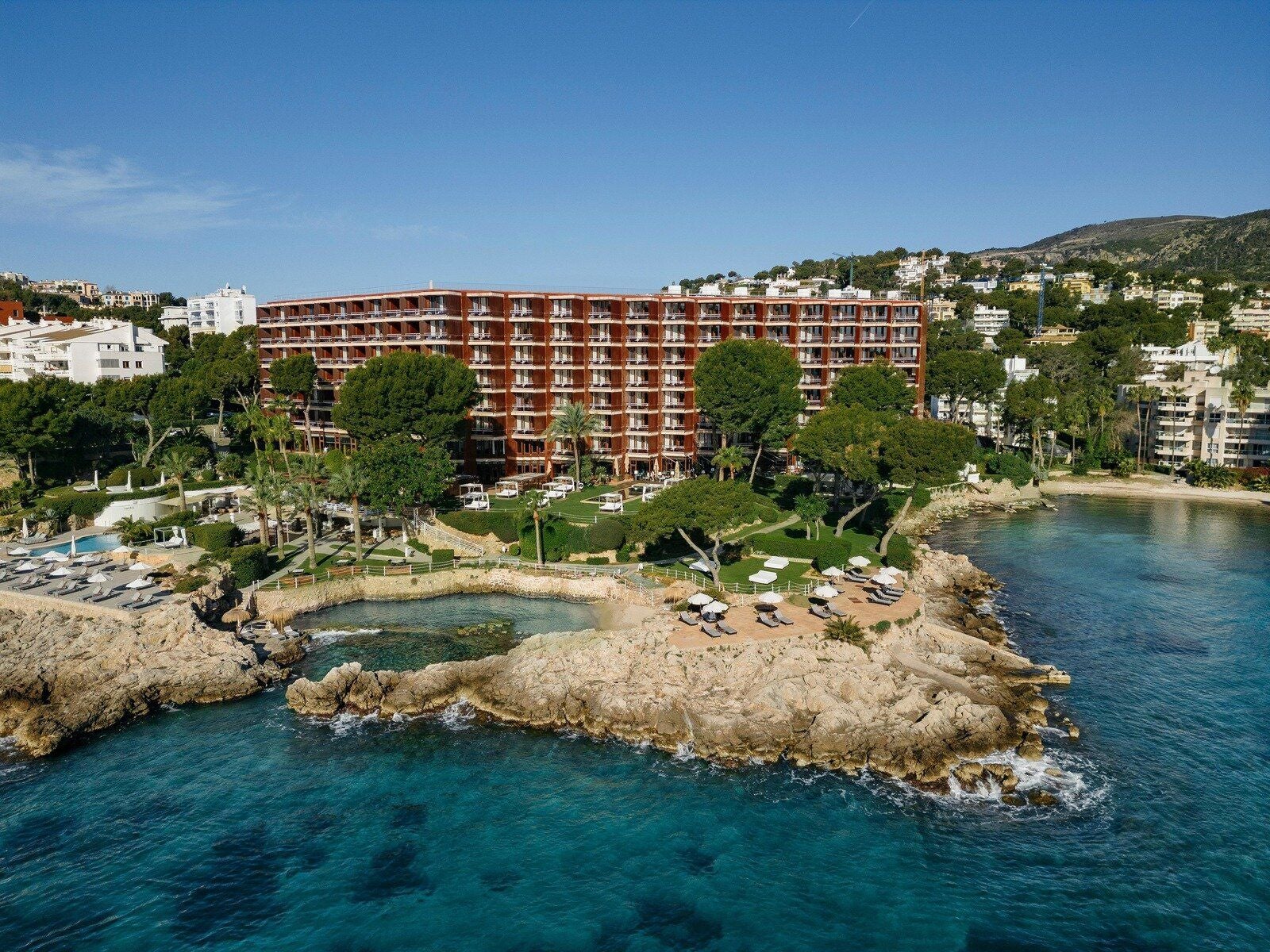 Großes Hotel am Meer mit Pool, Sonnenschirmen und üppigen Gärten an felsiger Küste.