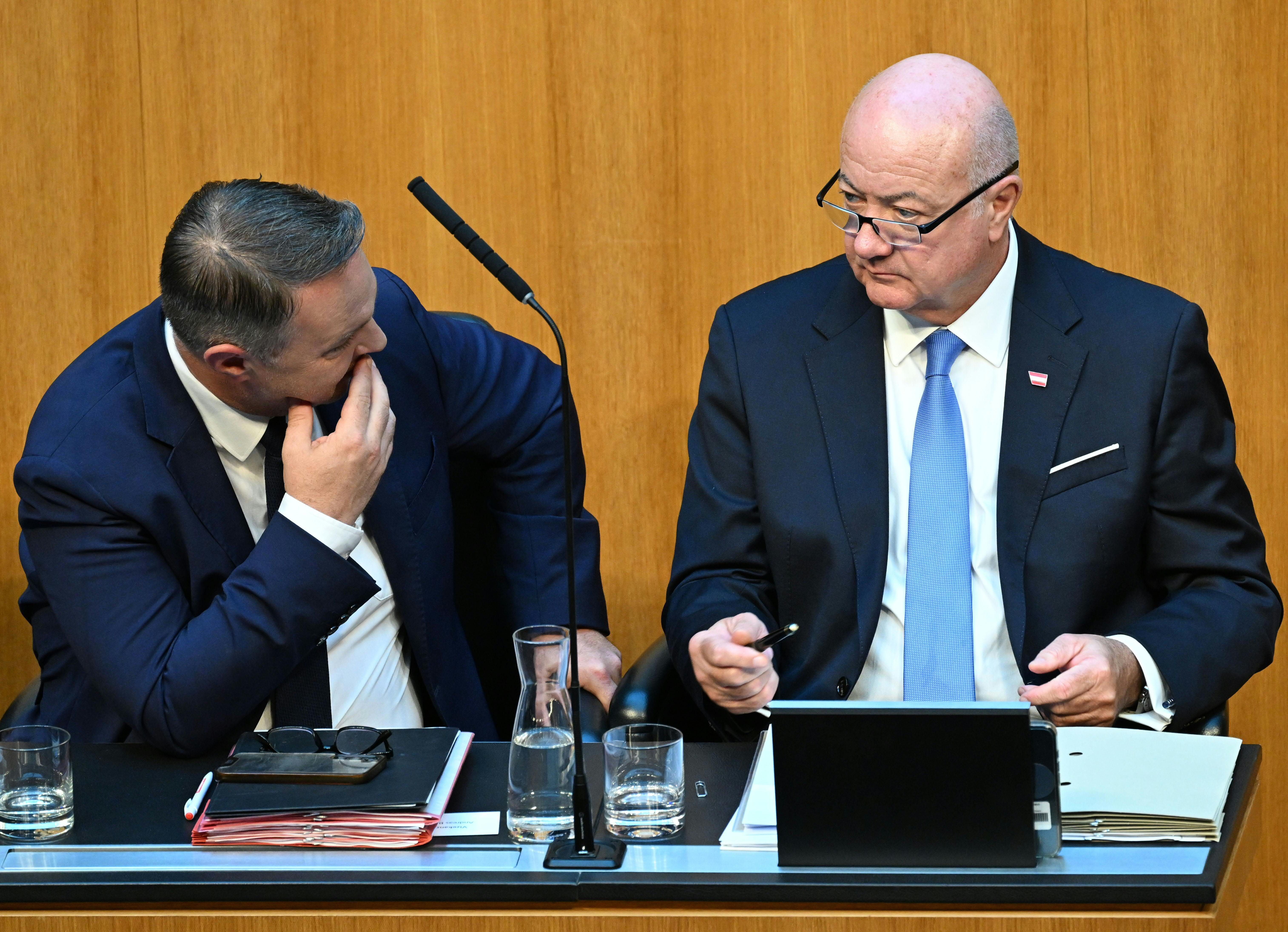 Zwei Männer im Anzug sprechen bei einer Parlamentssitzung am Tisch miteinander.