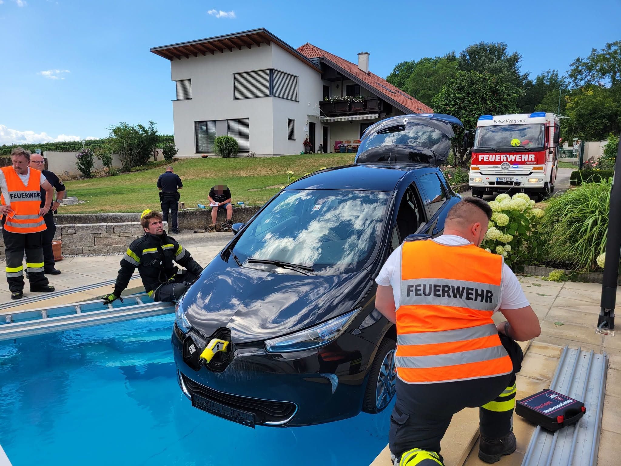 Mehrere Feuerwehrleute bergen ein Auto aus einem Pool vor einem Wohnhaus.